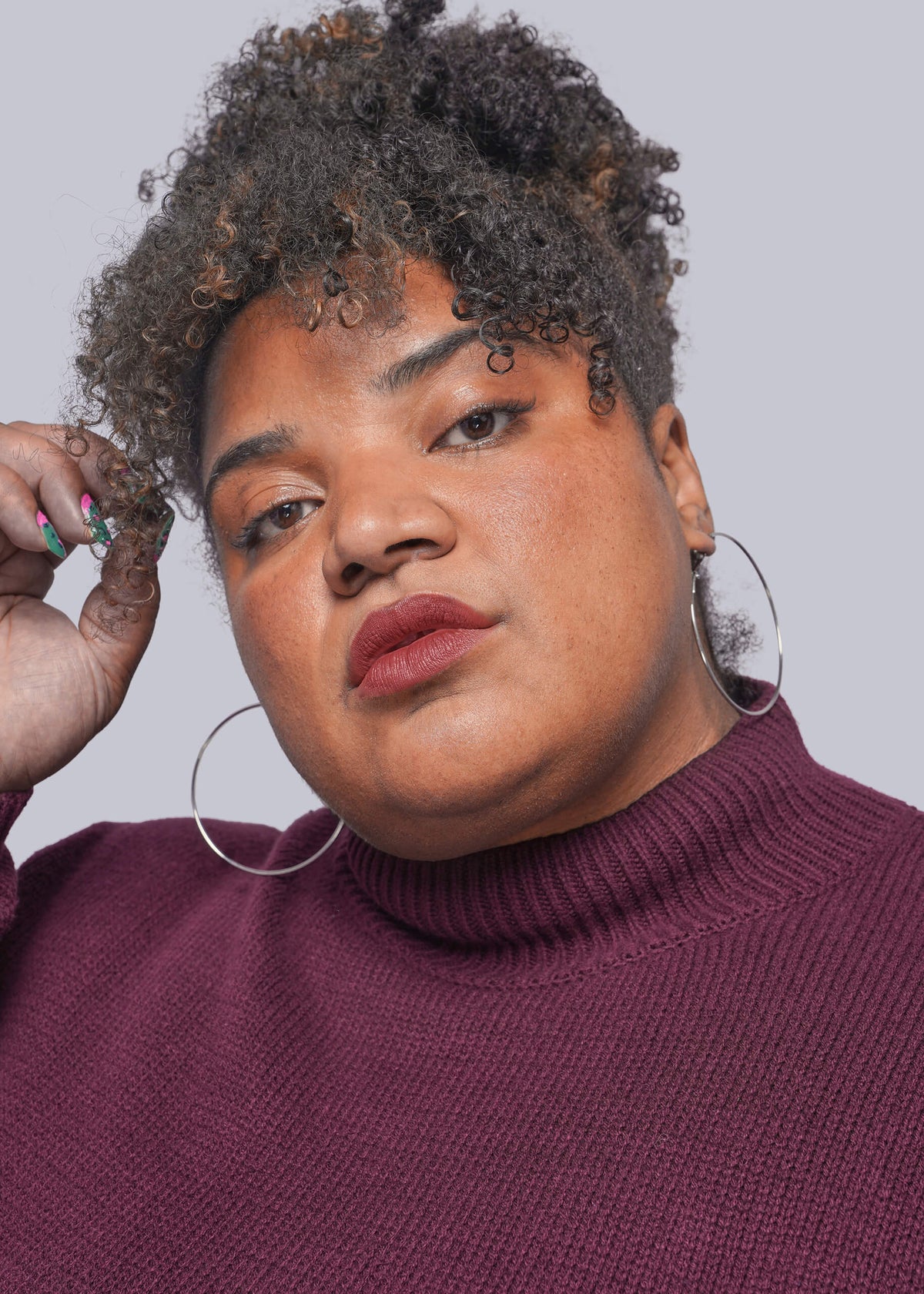 1X=A close-up portrait of a person with curly hair wearing a ribbed burgundy turtleneck sweater and large silver hoop earrings against a light gray studio background.