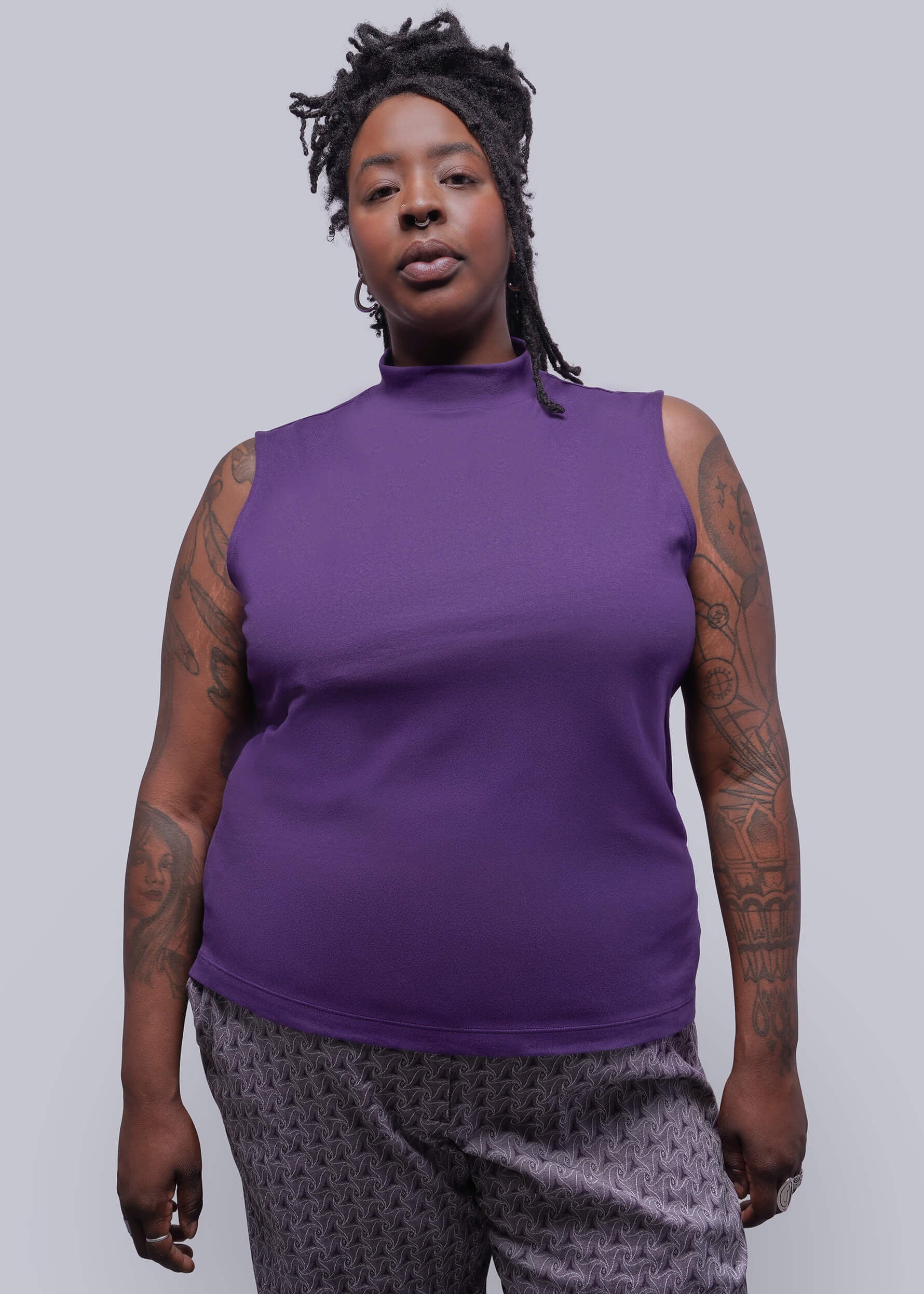 1X=Person wearing a purple sleeveless top and patterned pants against a plain background