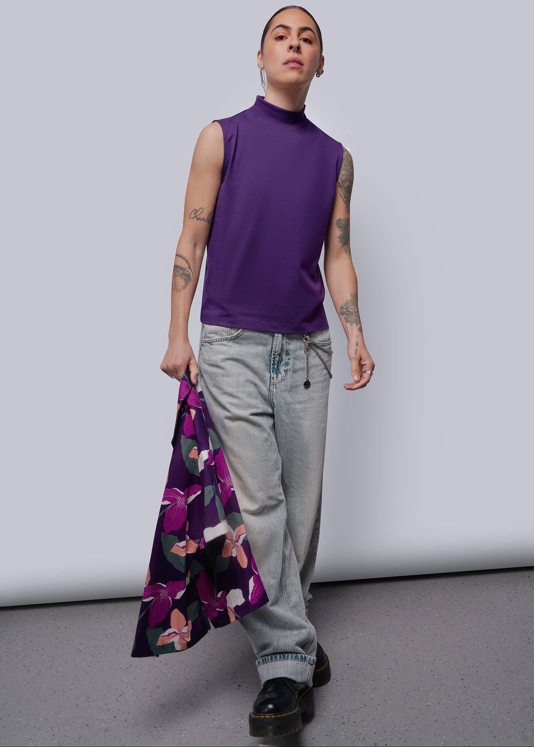 Person wearing a purple sleeveless top and gray pants, holding a floral shirt against a plain background.