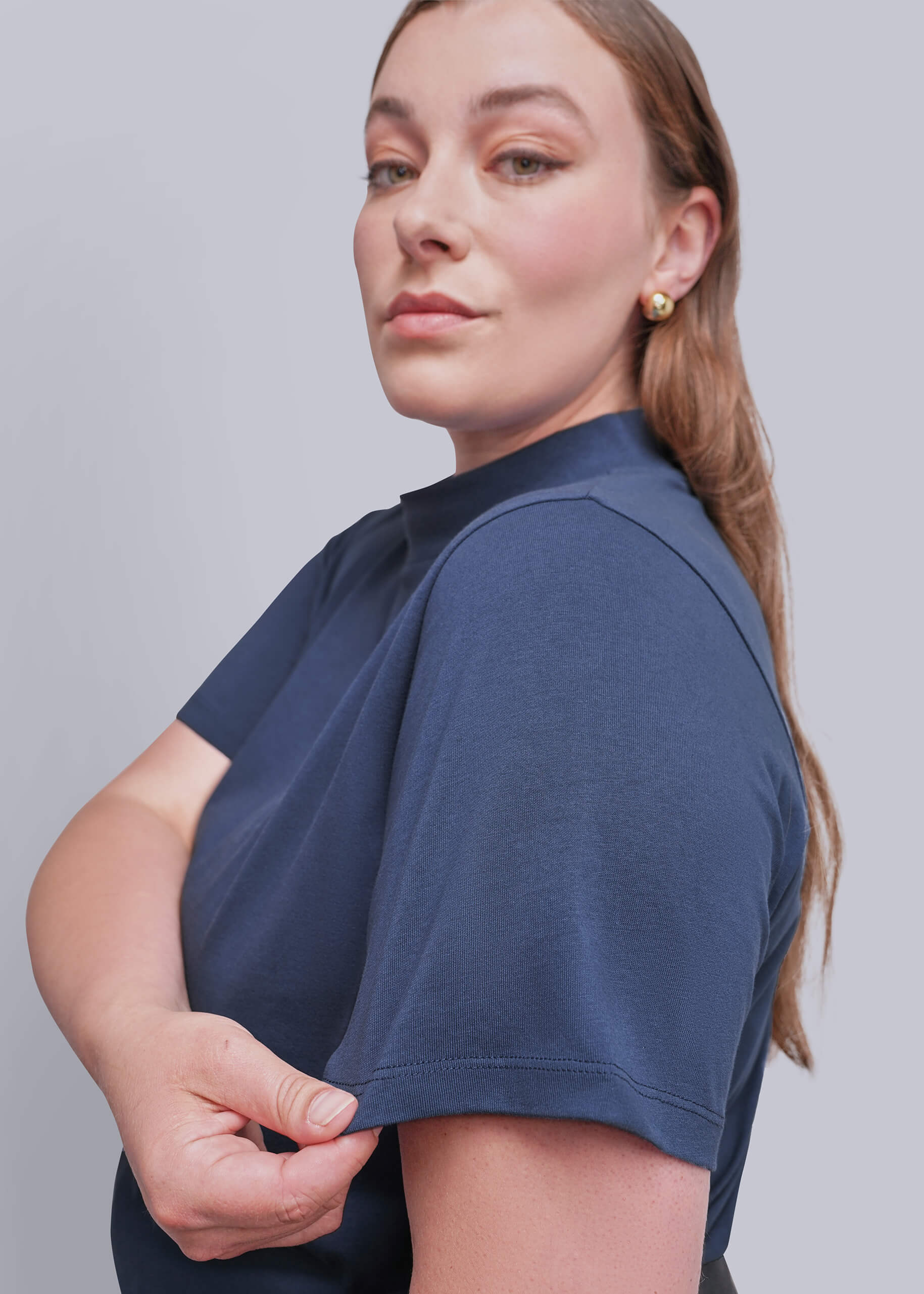 1X=A woman wearing a navy blue short-sleeve crewneck top, shown from a side-back angle against a solid light gray background.