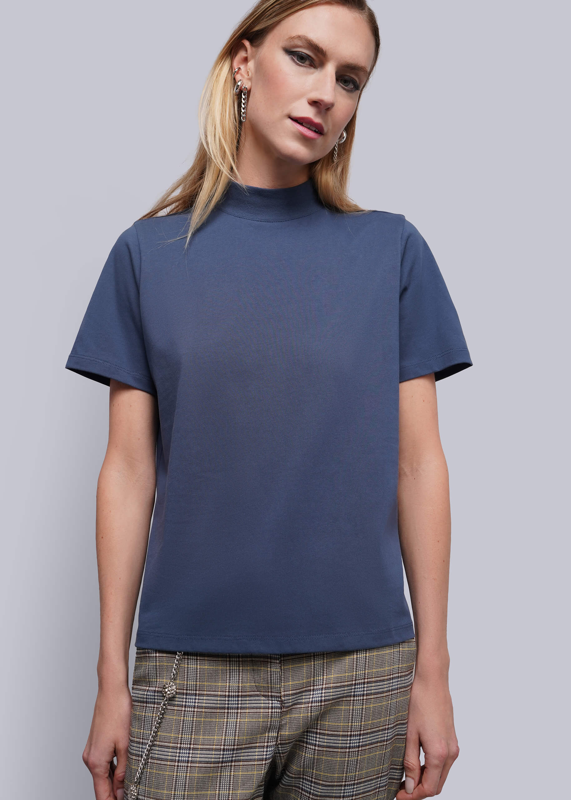 S=A woman wearing a short-sleeve mock neck t-shirt in slate blue, paired with plaid trousers, against a solid gray studio background.