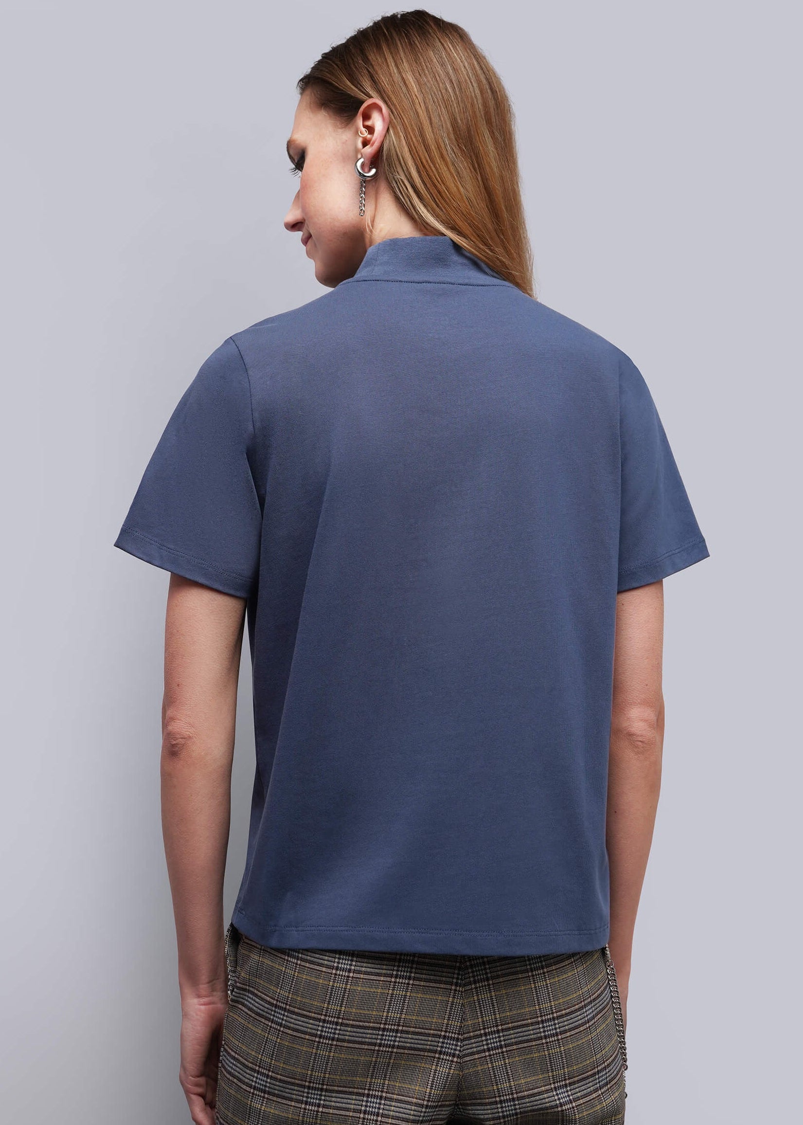 S=A woman shown from the back wearing a slate blue short-sleeve polo shirt and brown plaid trousers, photographed against a solid light gray studio background.