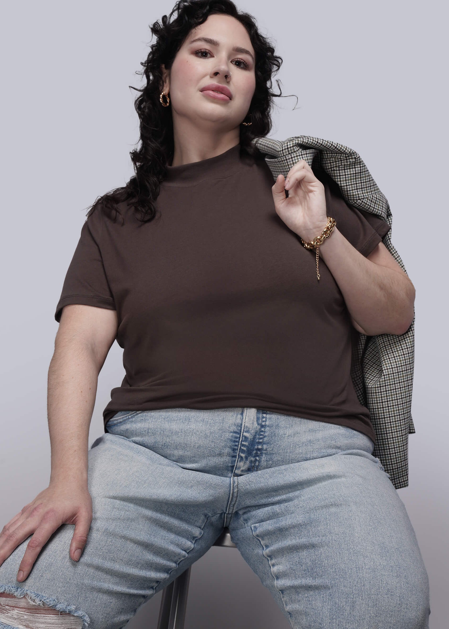 1X=Person wearing a brown t-shirt and light blue jeans, holding a plaid jacket against a plain background