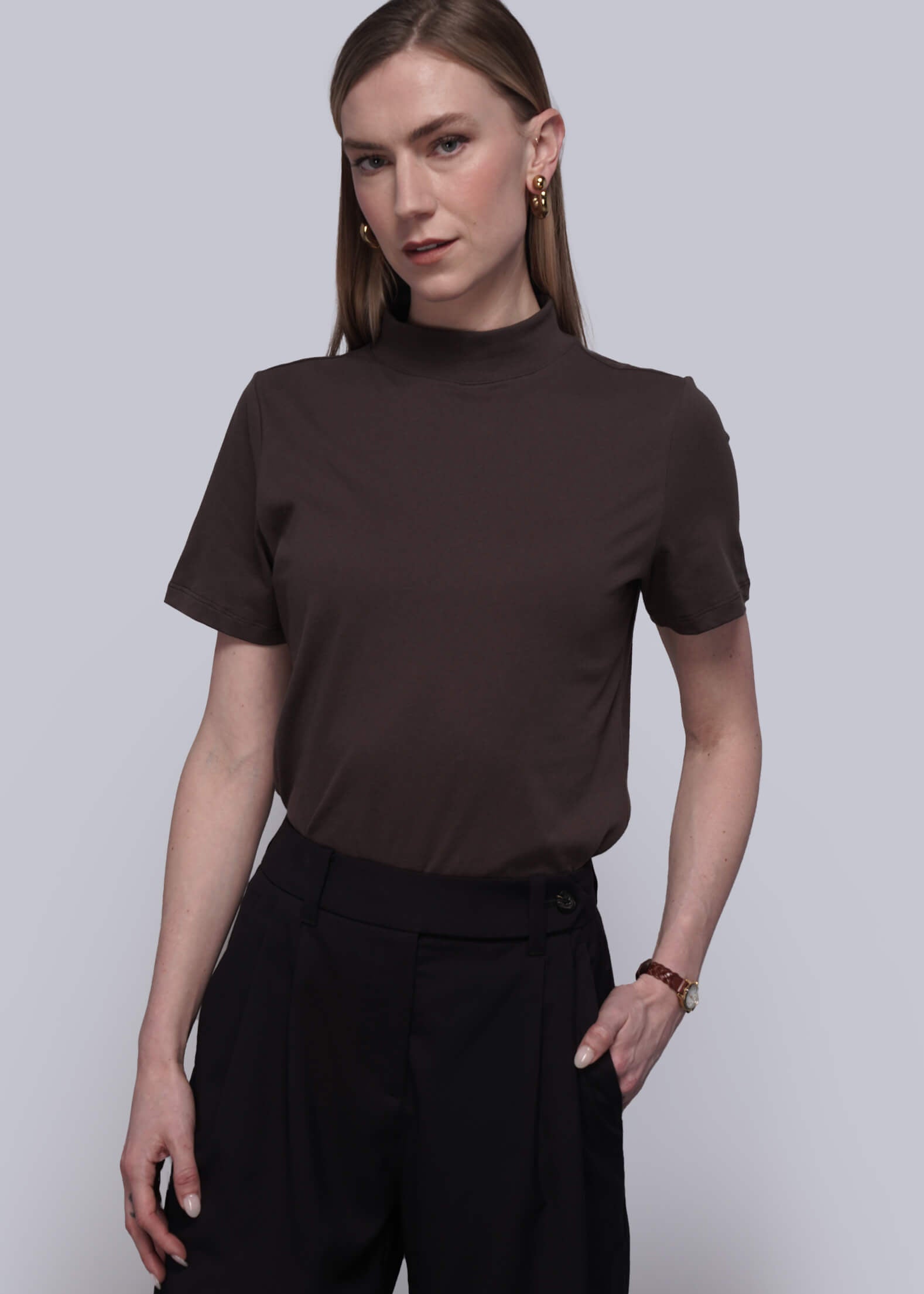 S=Woman wearing a brown t-shirt and black pants against a light gray background