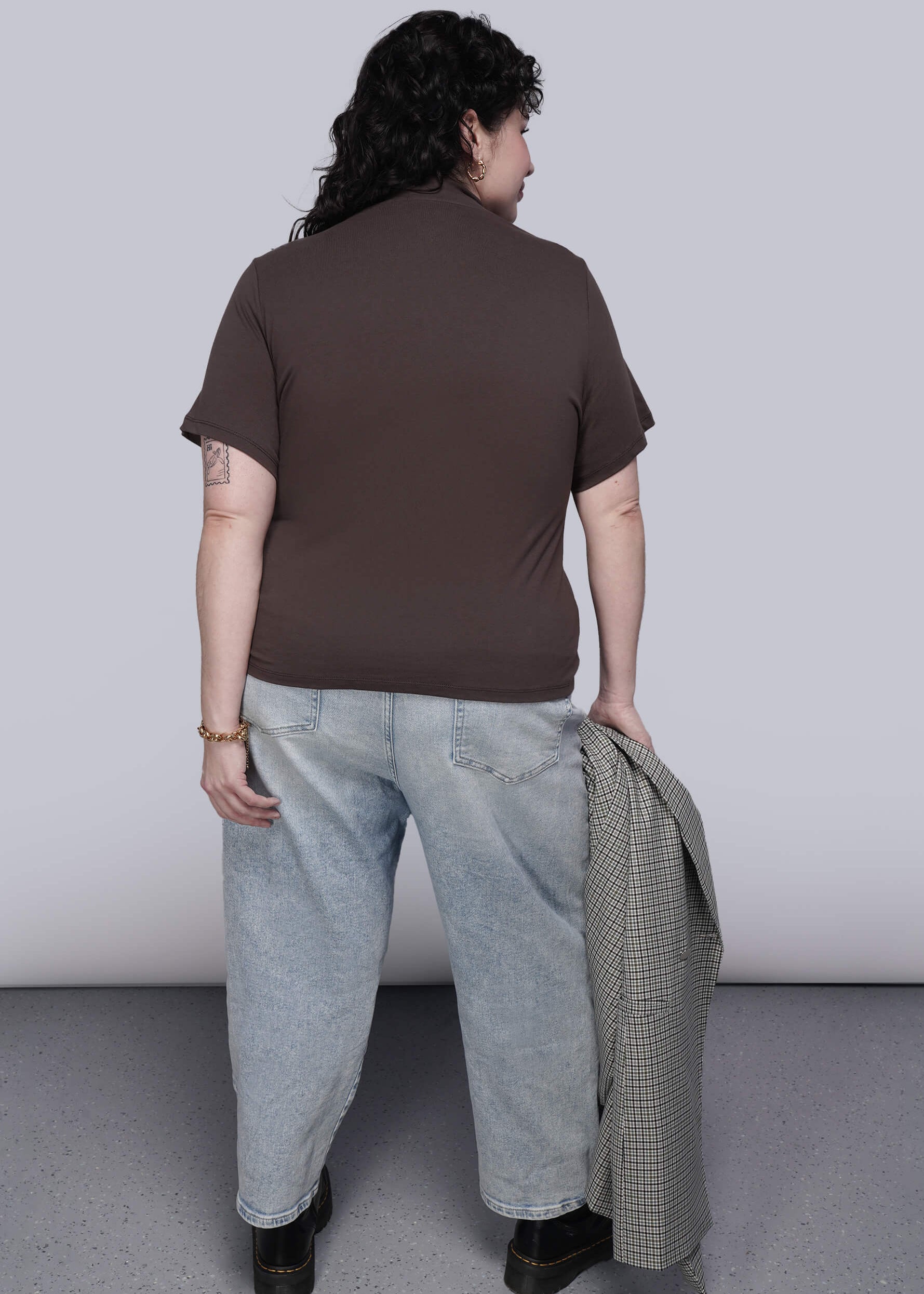 1X=Back of person wearing a brown shirt and light blue jeans, holding a plaid jacket against a plain background