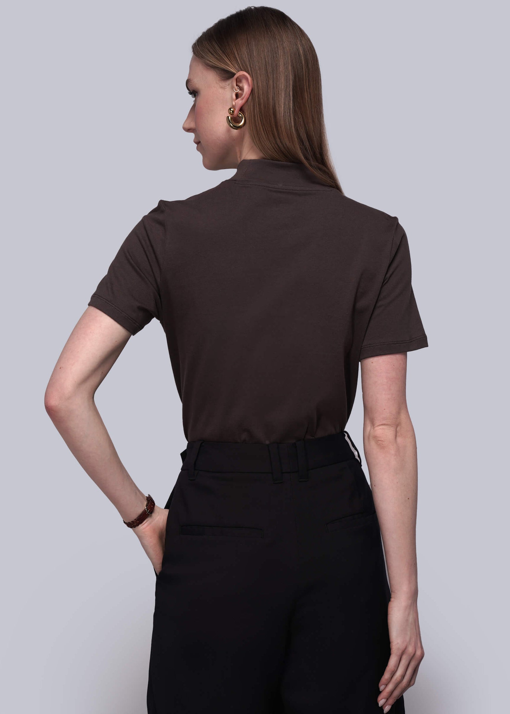 S=Woman wearing a brown short-sleeve shirt and black pants against a light gray background