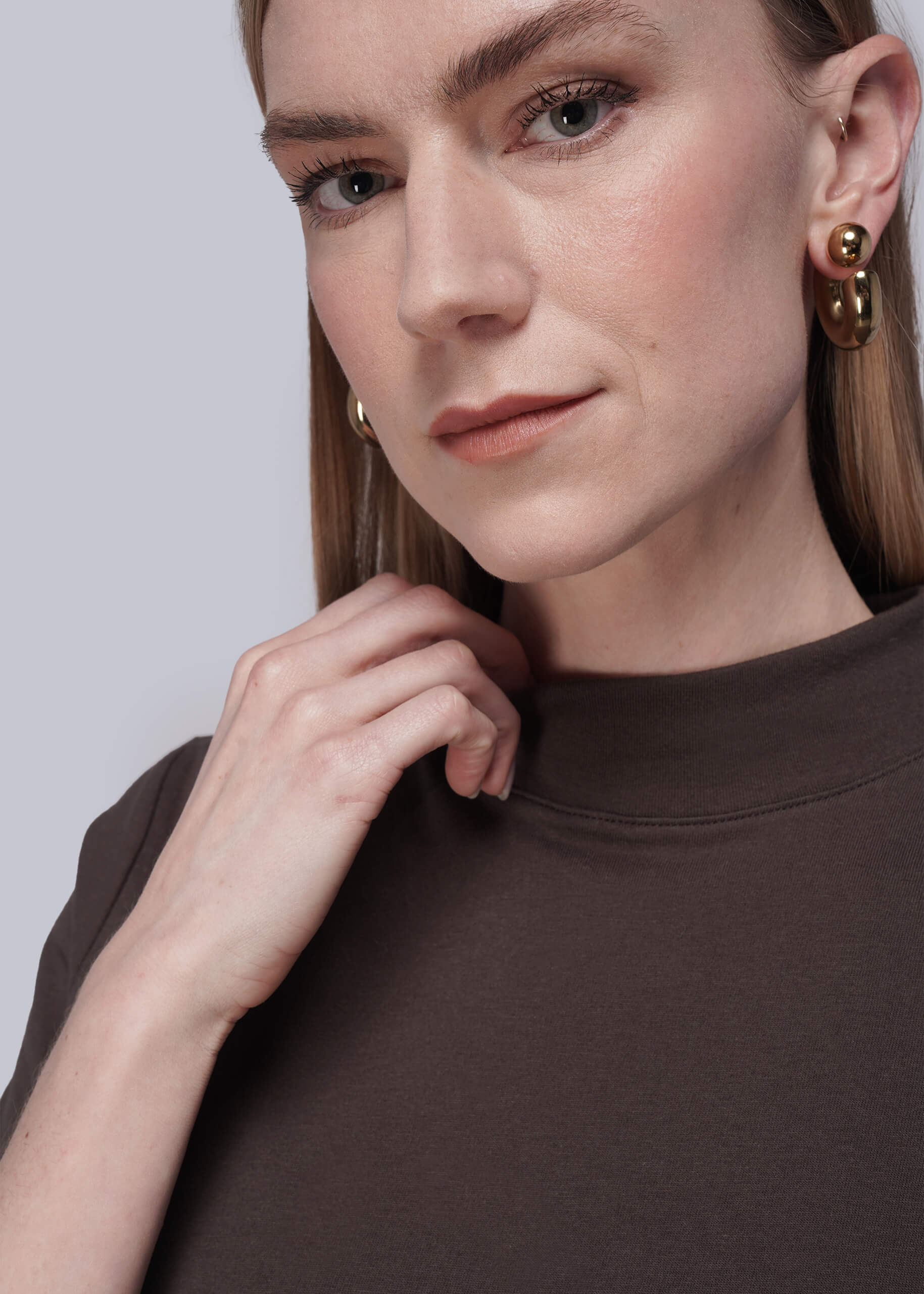 S=Close-up of a person wearing brown shirt and gold earrings against a light gray background
