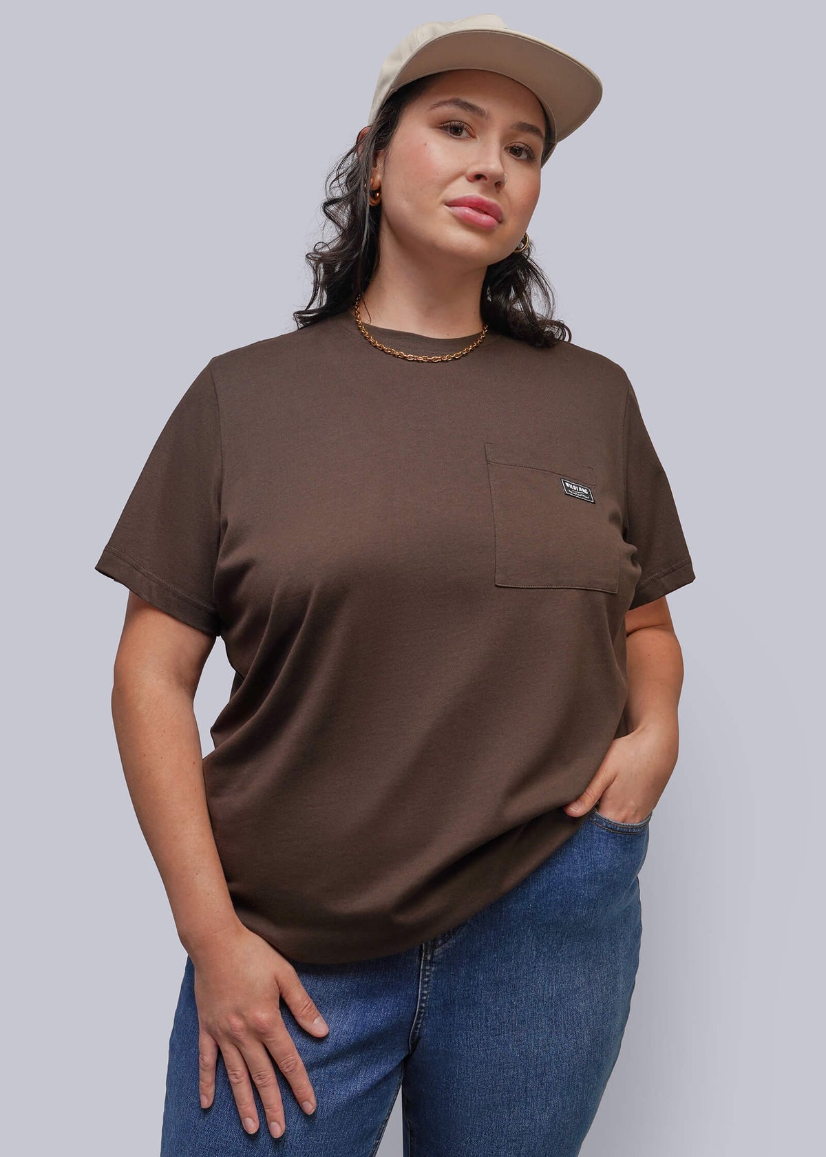 1X=A woman modeling a brown short-sleeve crewneck t-shirt with a chest pocket, paired with blue jeans and a beige baseball cap, against a solid gray background.
