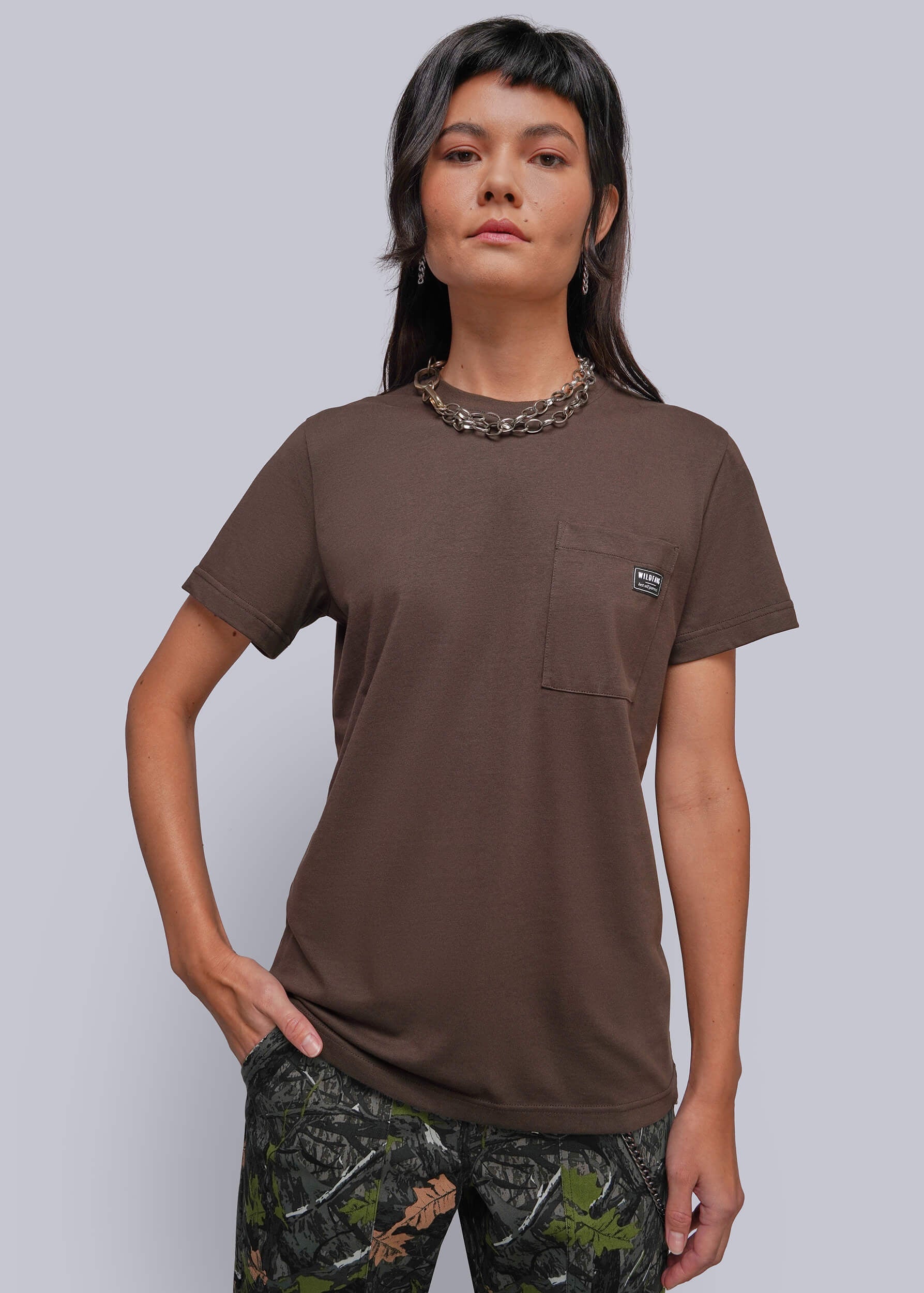 A woman modeling a brown short-sleeve pocket t-shirt with a small embroidered logo on the chest pocket, paired with camouflage pants, against a solid gray studio background.