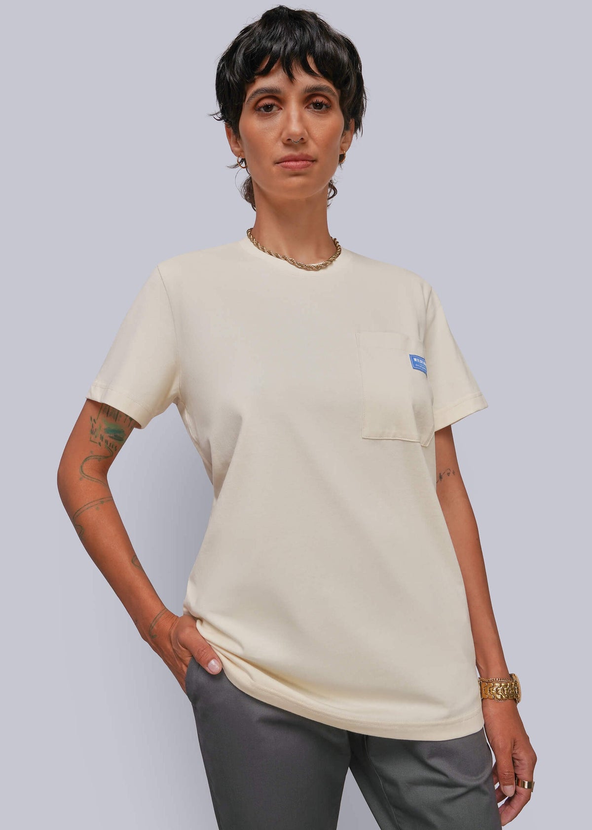 S=A model wearing a cream-colored short-sleeve pocket t-shirt with a small blue logo on the chest pocket, paired with gray pants, against a solid light gray background.