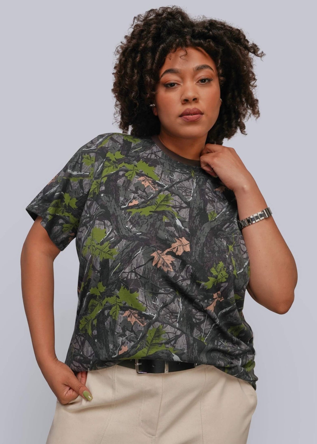 1X=A woman modeling a short-sleeve crewneck t-shirt with a dark gray and green woodland camouflage pattern, paired with beige pants, against a solid light gray background.