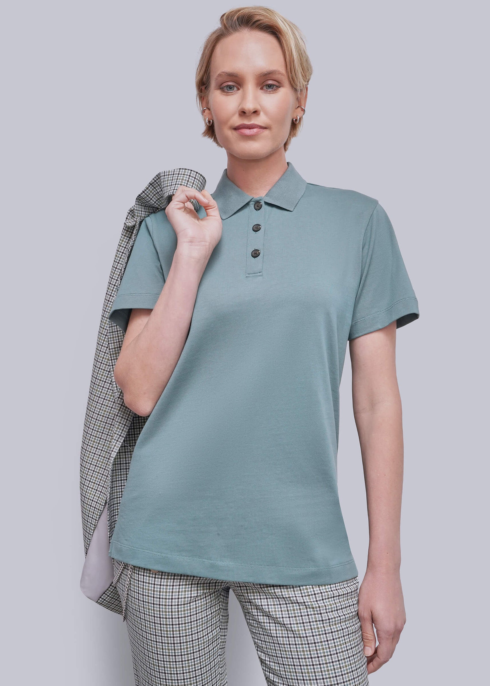 A woman modeling a short-sleeve polo shirt in a muted teal color, paired with matching plaid trousers and holding a plaid jacket over her shoulder, against a solid gray studio background.