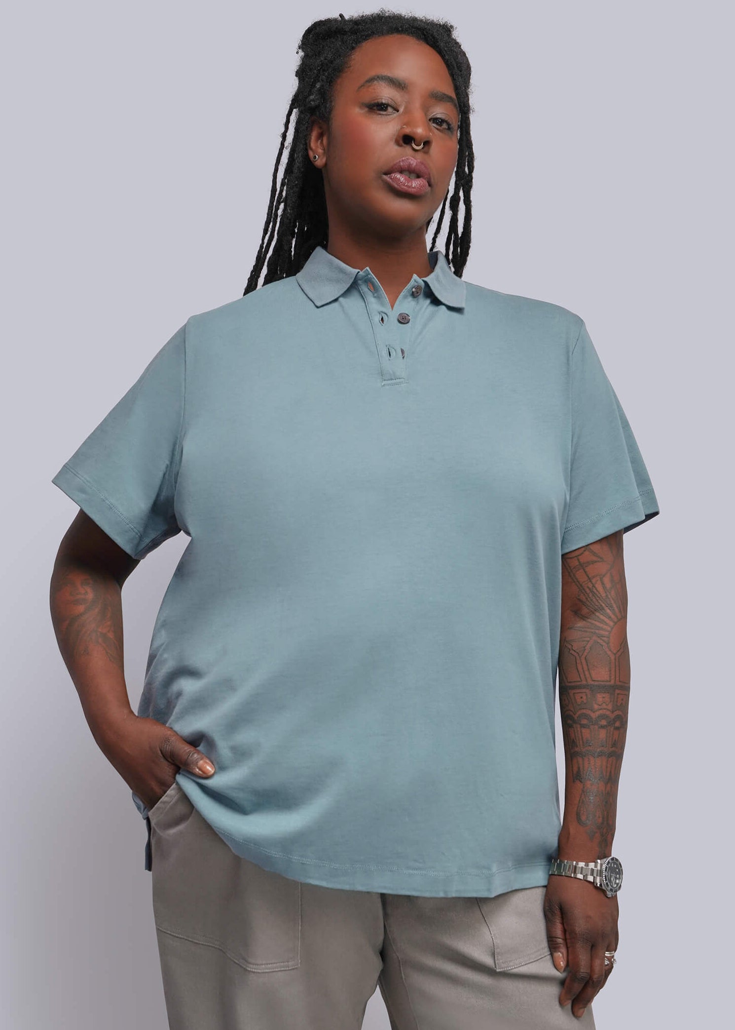 1X=A woman modeling a light blue short-sleeve polo shirt with beige trousers, standing against a solid light gray studio background.