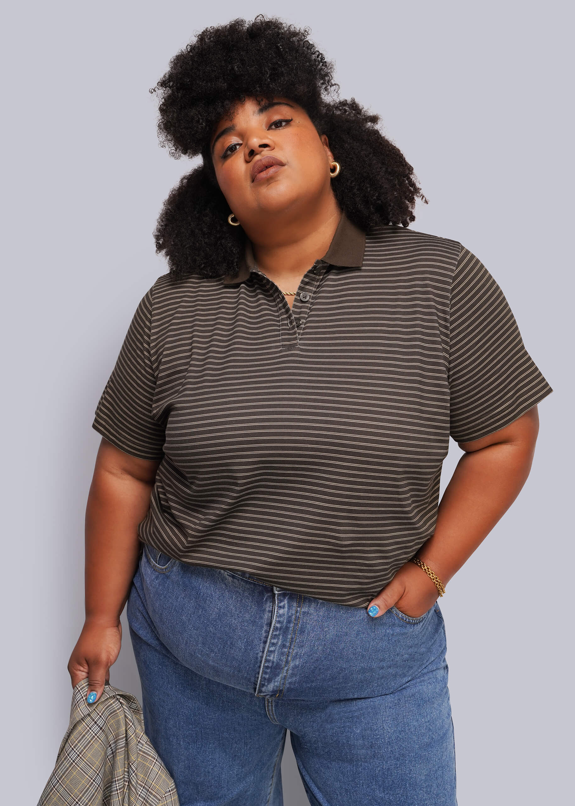 1X=A plus-size model wearing a dark brown and black horizontally striped short-sleeve polo shirt and blue jeans, holding a plaid shirt in one hand against a solid gray studio background.