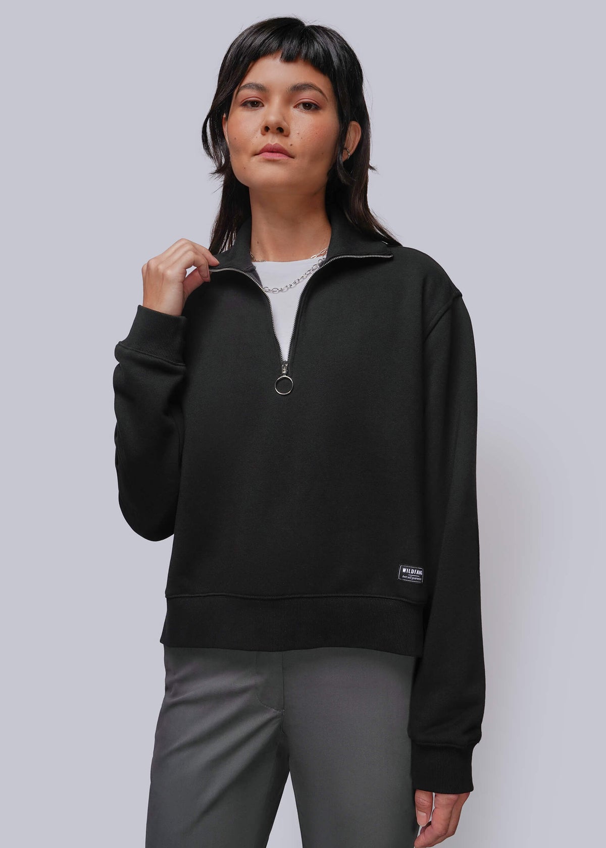 S=A woman modeling a black half-zip sweatshirt with a small circular zipper pull and a small black label on the lower left hem, paired with gray pants against a light gray studio background.
