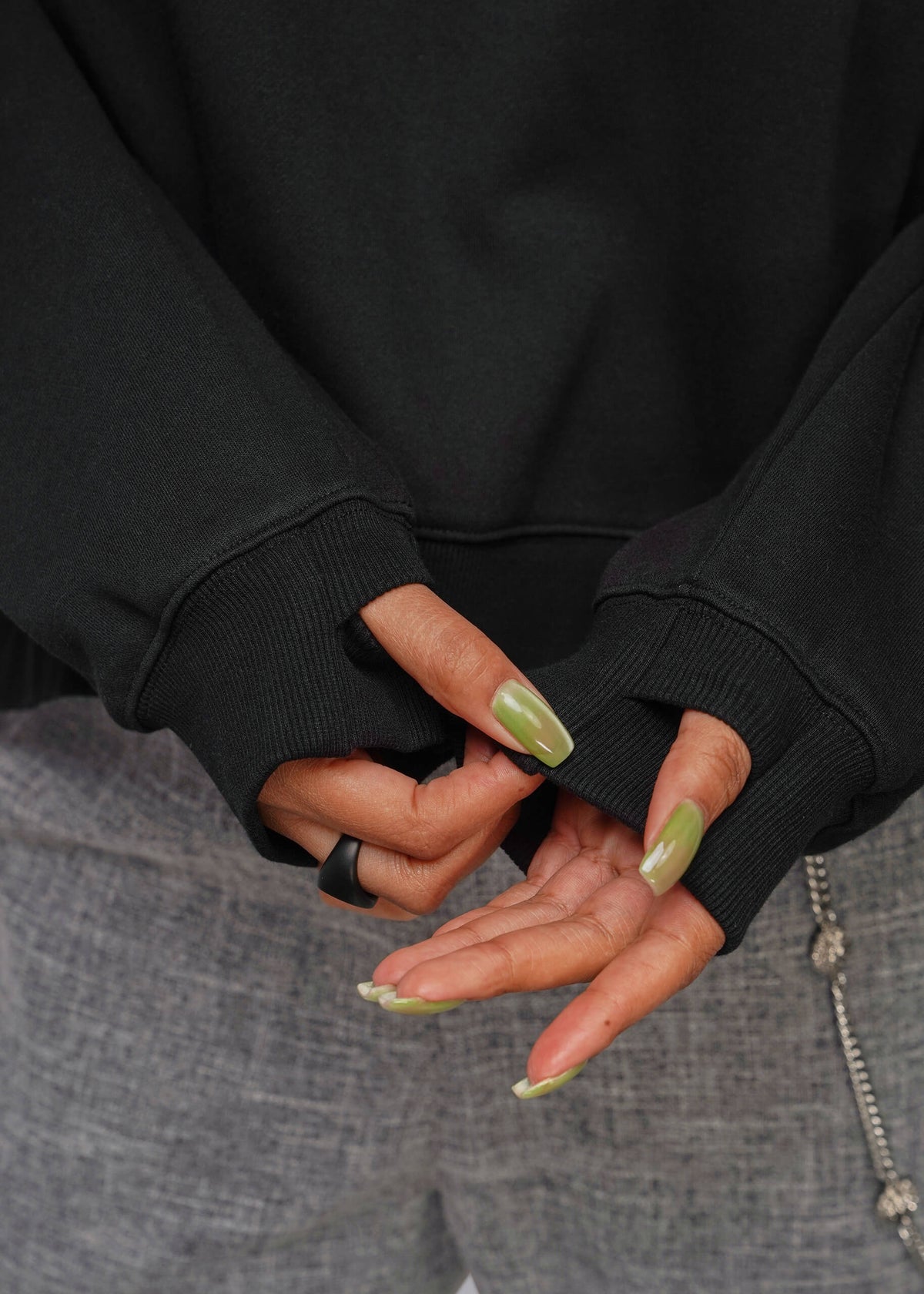 1X=Close-up of a person wearing a sweatshirt with thumbholes in the sleeces, showing manicured nails with a metallic olive green glitter polish and a black ring on one finger, paired with gray textured pants.