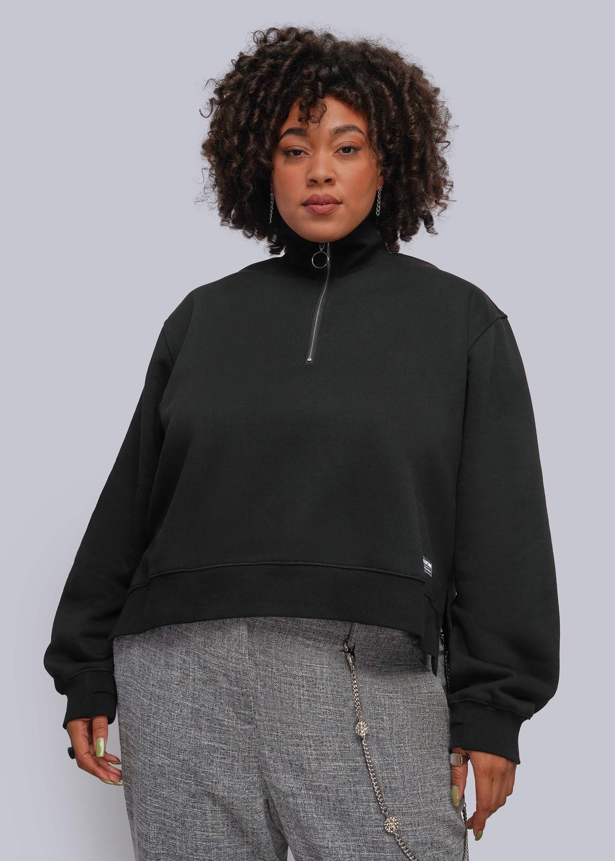 1X=A model with curly dark hair wearing a black cropped quarter-zip sweatshirt and gray textured pants, standing against a solid light gray studio background.