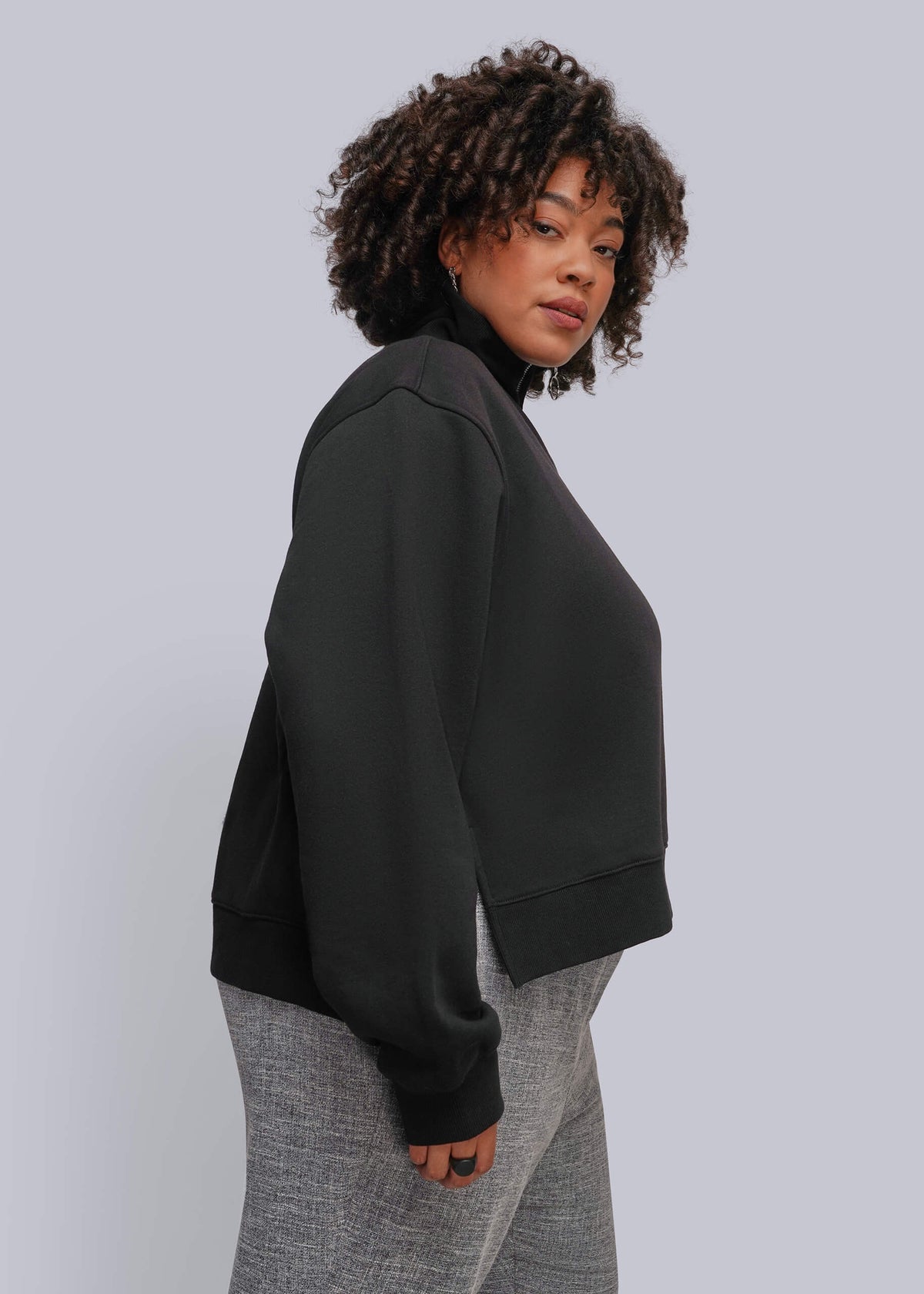 1X=A woman with curly dark hair is shown from a side-back angle wearing a black cropped crewneck sweatshirt and gray textured pants against a solid light gray studio background.