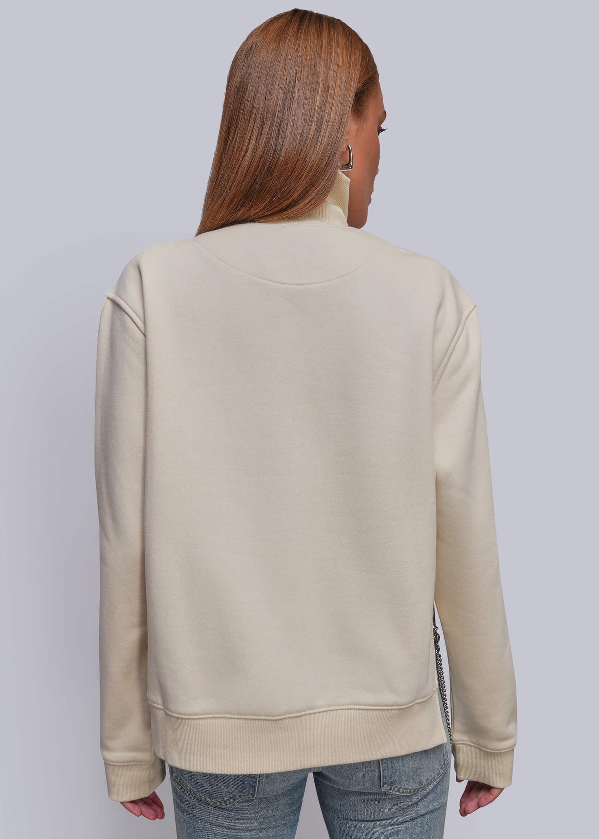 S=A woman viewed from the back wearing a light beige sweatshirt and blue jeans against a solid light gray studio background.
