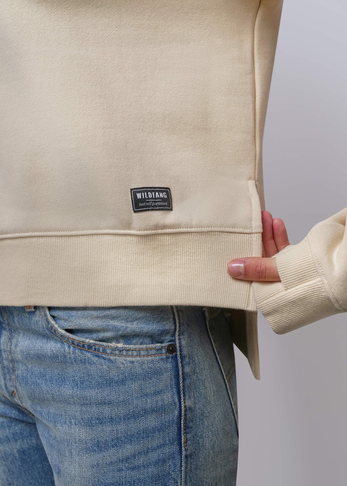S=Close-up of a cream-colored sweatshirt with a ribbed hem and a black rectangular 'WILDFANG' brand patch, worn by a model with blue jeans.