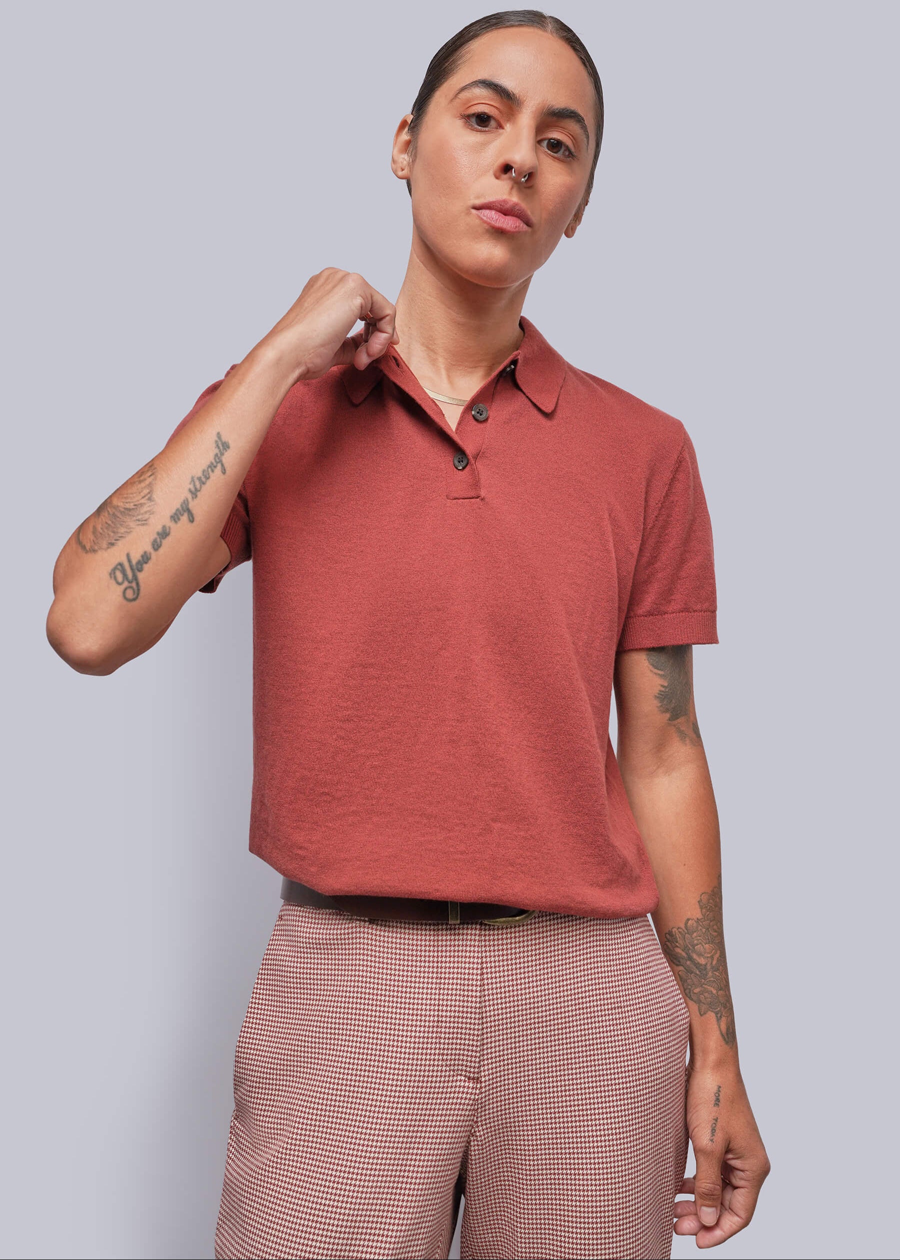 S=A model wearing a short-sleeve polo shirt in a muted terracotta or dusty rose-red color, paired with red and white houndstooth trousers, against a solid light gray background.