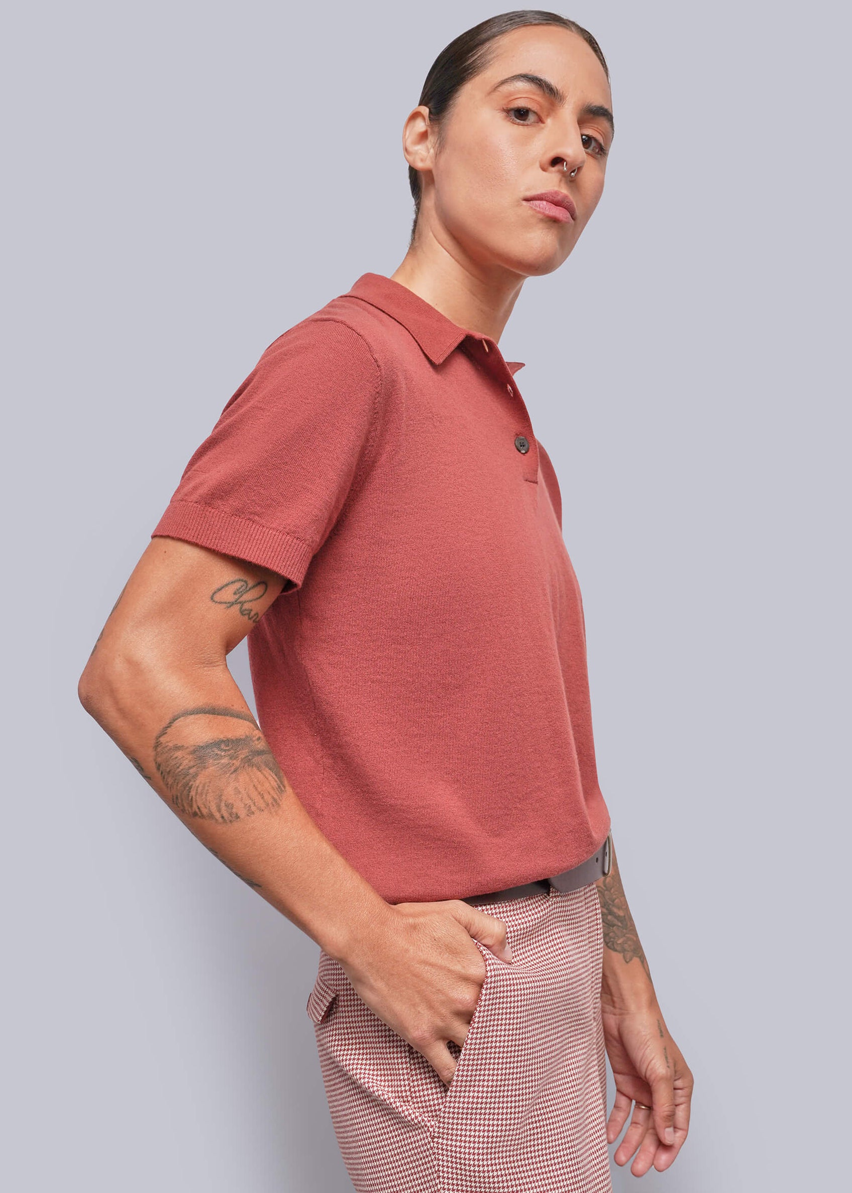 S=A person wearing a muted red short-sleeve polo shirt and red-and-white houndstooth trousers, photographed against a solid light gray studio background.
