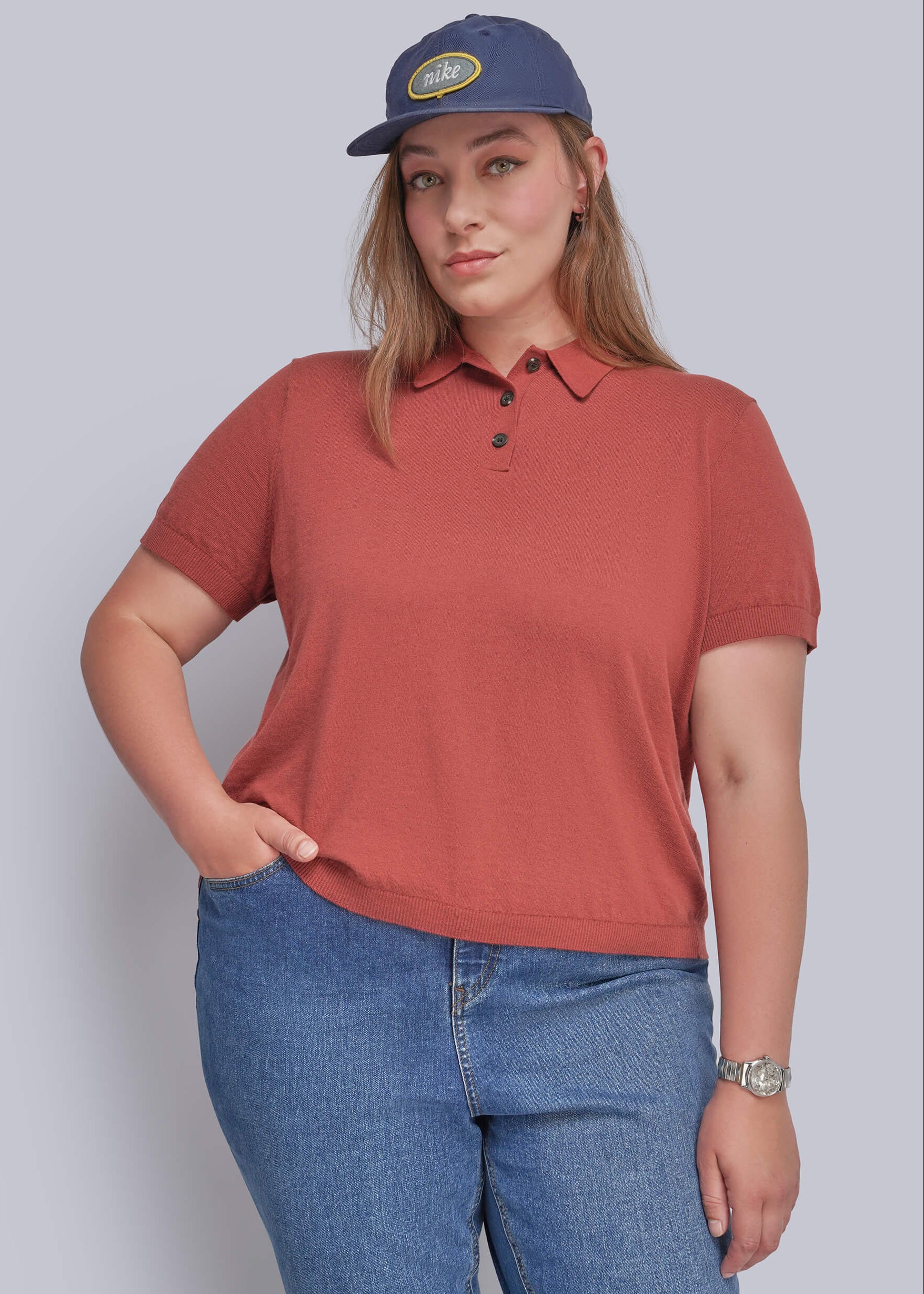 1X=A woman modeling a short-sleeve polo shirt in a muted terracotta or rust color, paired with blue jeans and a navy blue baseball cap, against a solid light gray background.