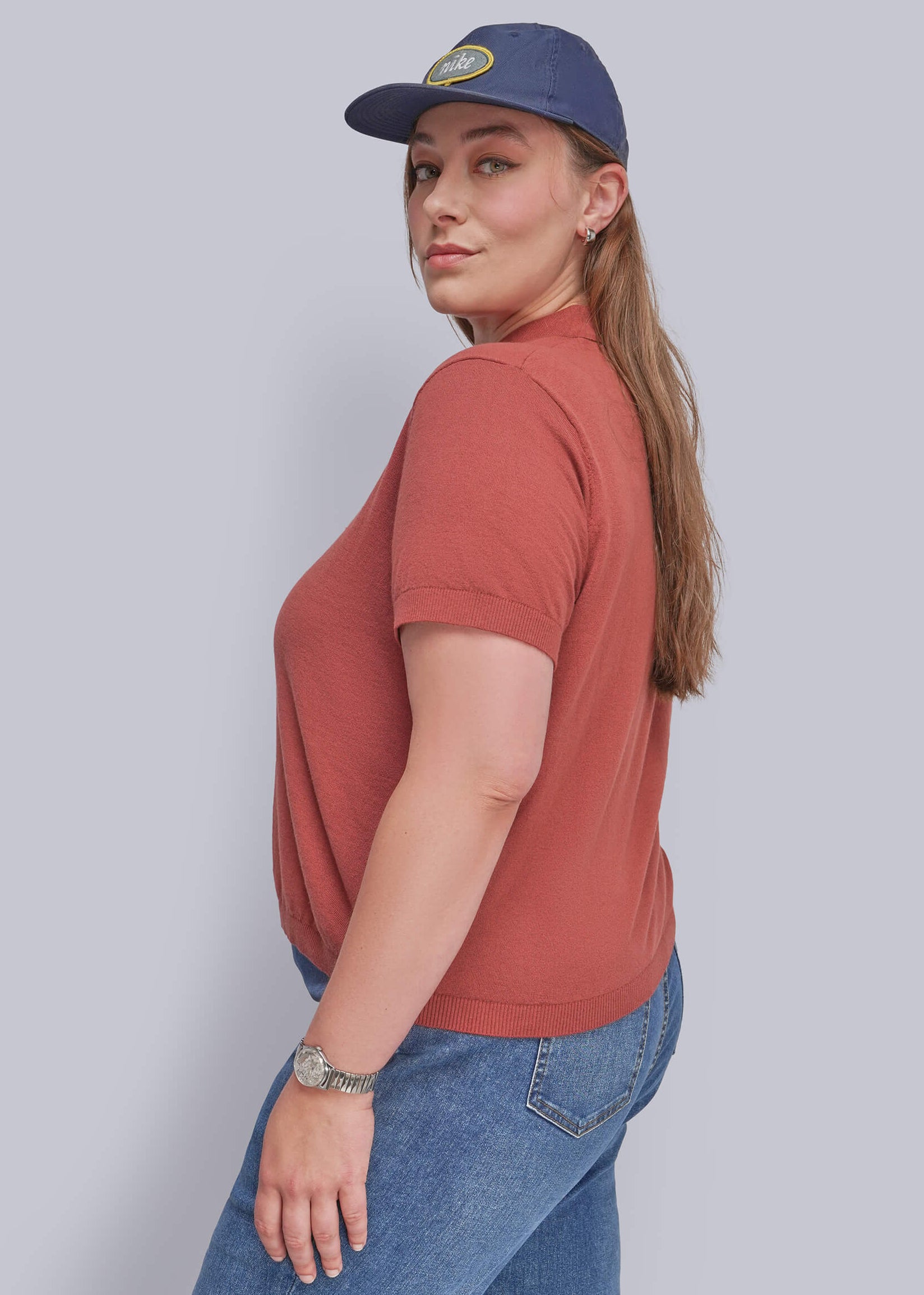 1X=A woman wearing a rust-colored short-sleeve t-shirt, blue jeans, and a navy blue baseball cap, shown from a side-back angle against a solid gray studio background.