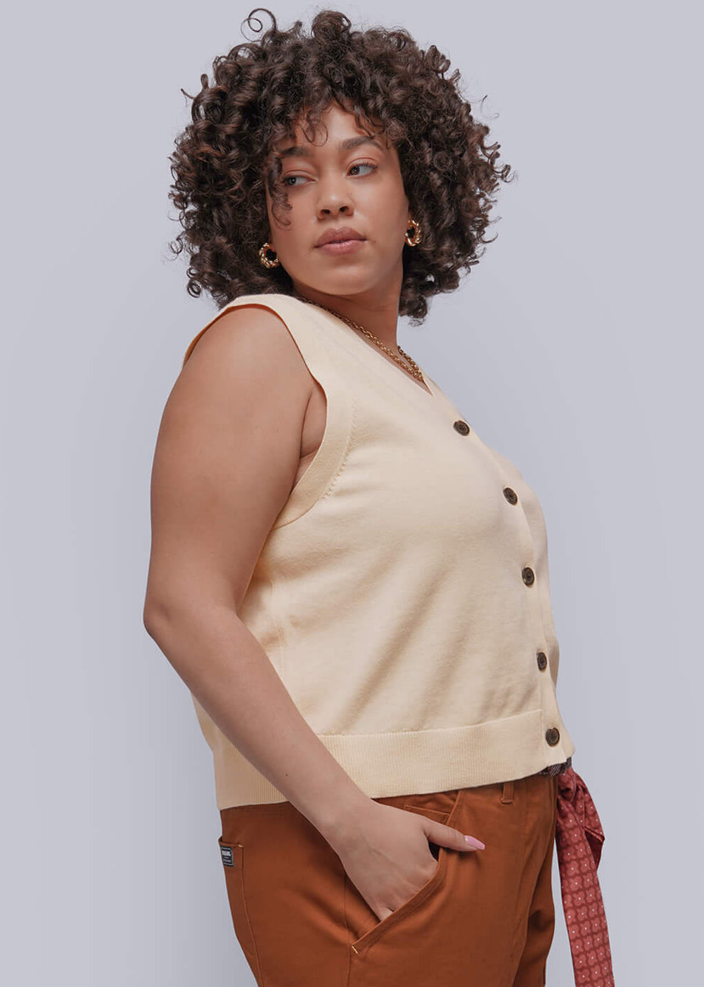 1X=Woman wearing a beige sweater vest  and brown pants against a white background