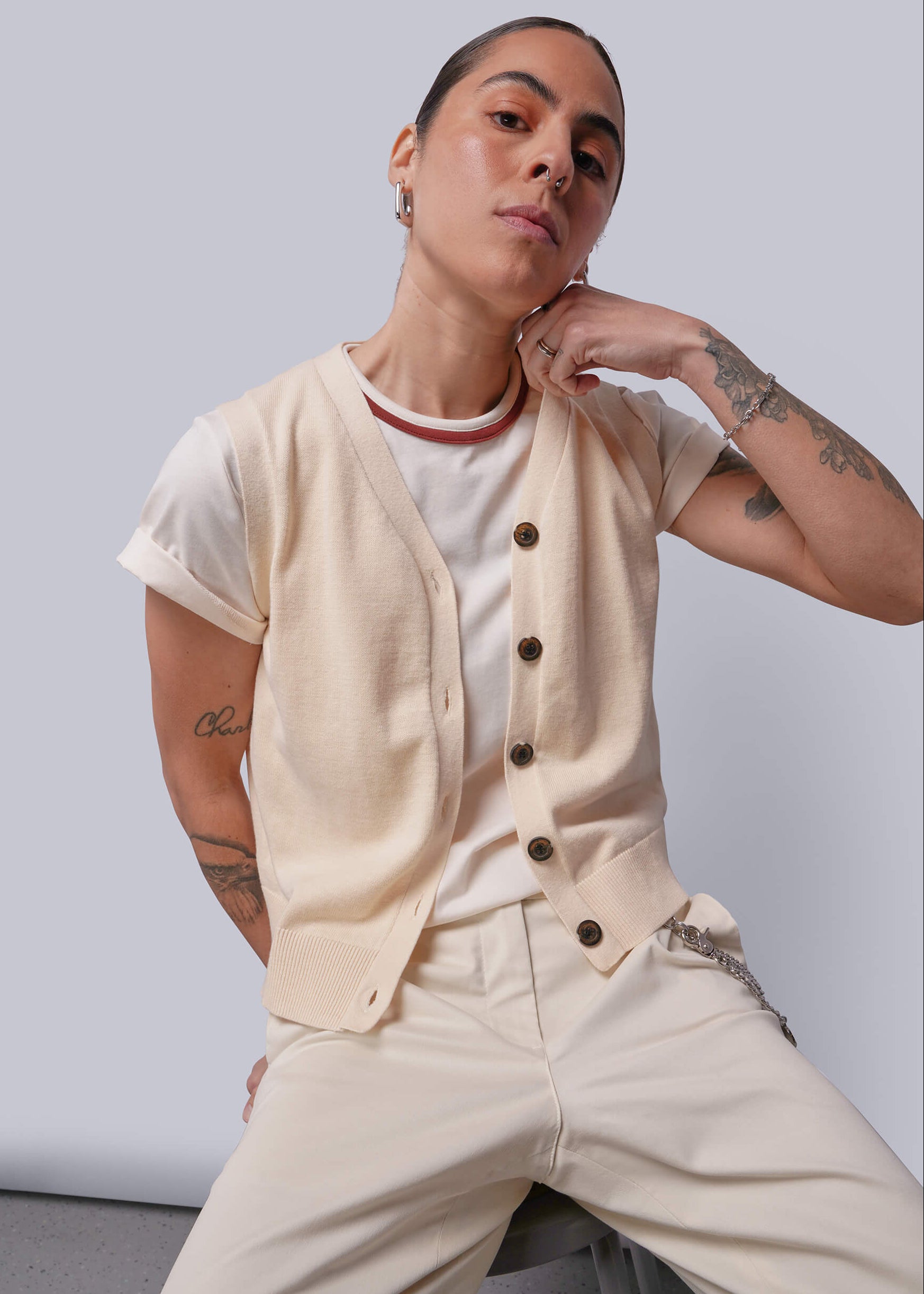 Person wearing a beige sweater vest over a white shirt with beige pants against a plain background