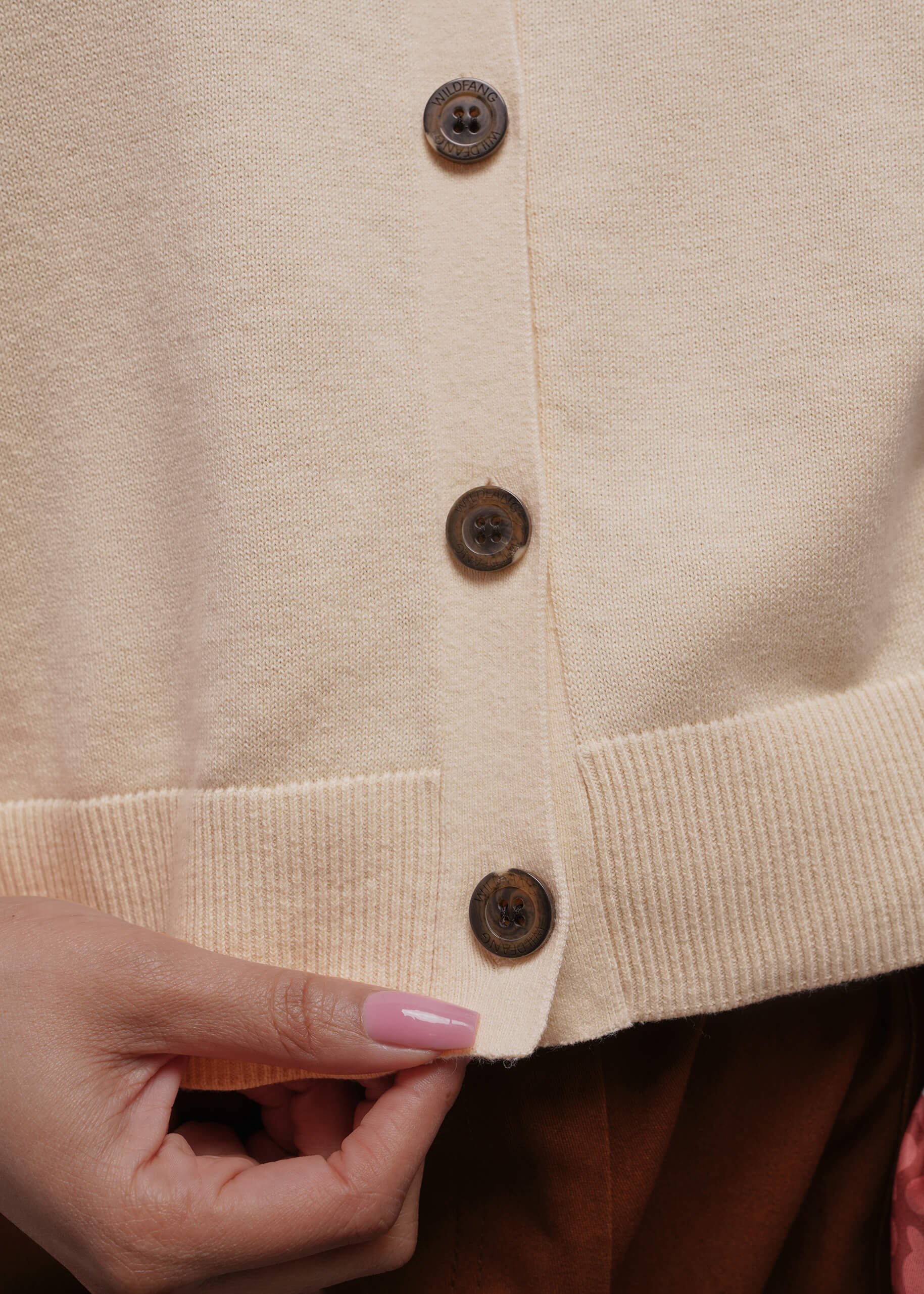 1X=Close-up of a beige sweater vest with buttons and a hand adjusting it.