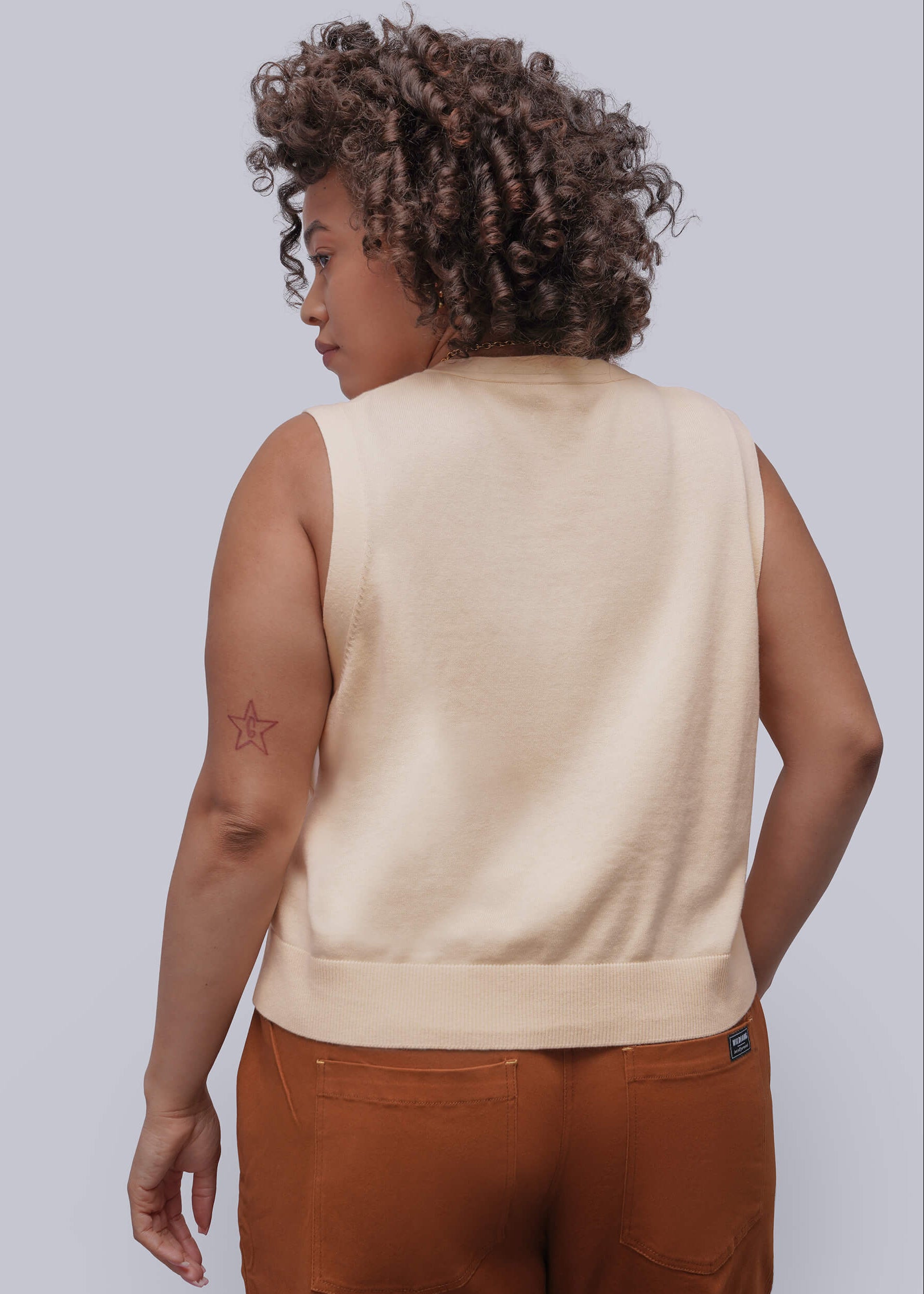 1X=Person wearing a beige sweater vest and brown pants against a light gray background
