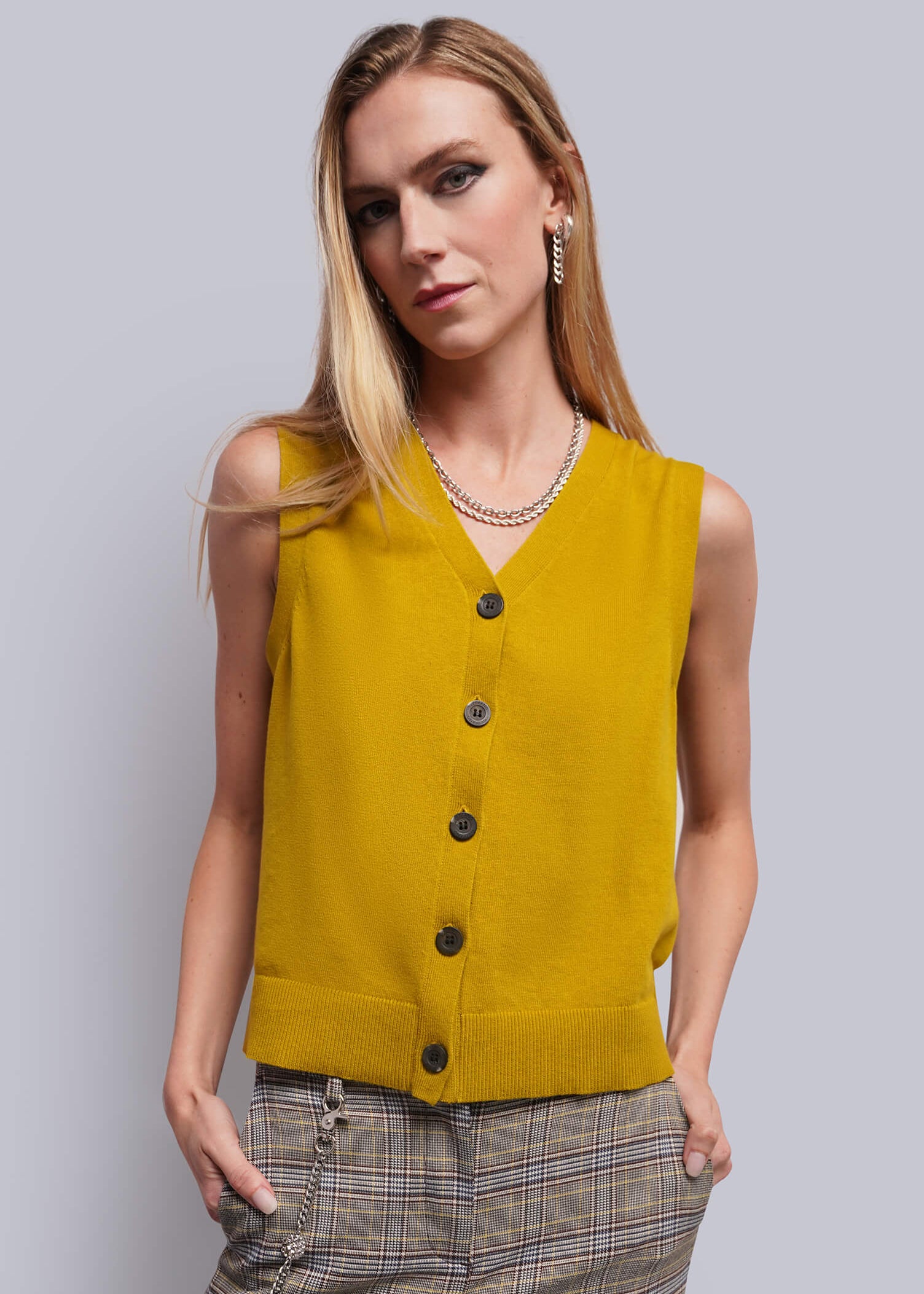 A woman modeling a sleeveless mustard yellow knit vest with dark buttons, paired with brown plaid trousers, against a solid gray studio background.