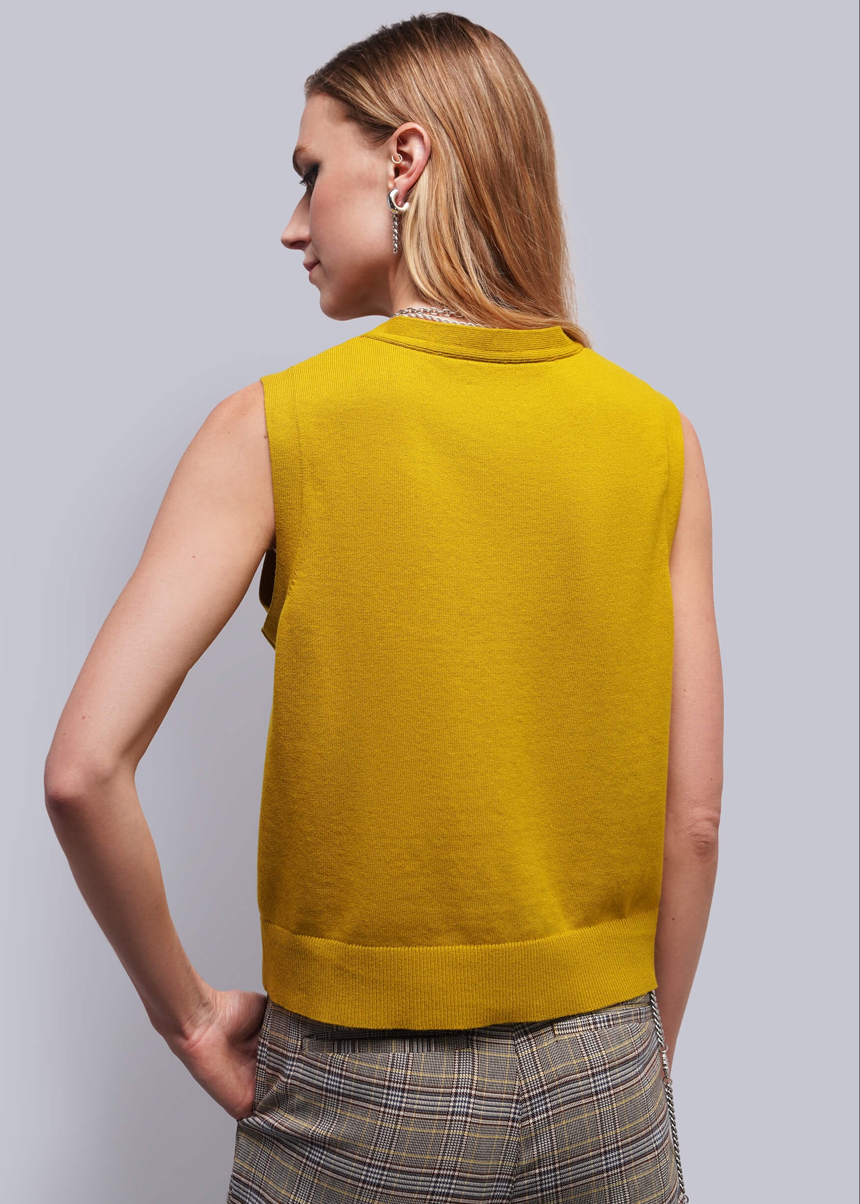 S=A woman shown from the back wearing a sleeveless mustard yellow knit top and plaid trousers, photographed against a plain light gray studio background.