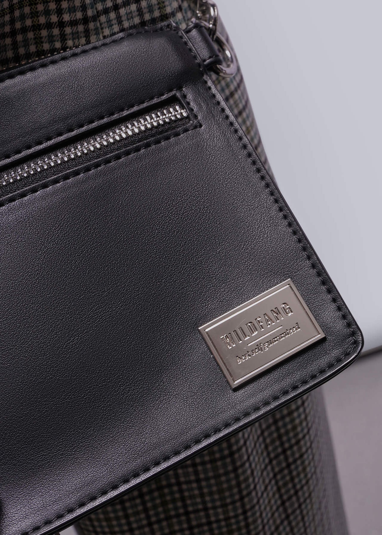 Black leather bag with a brand logo held by a person wearing plaid pants.
