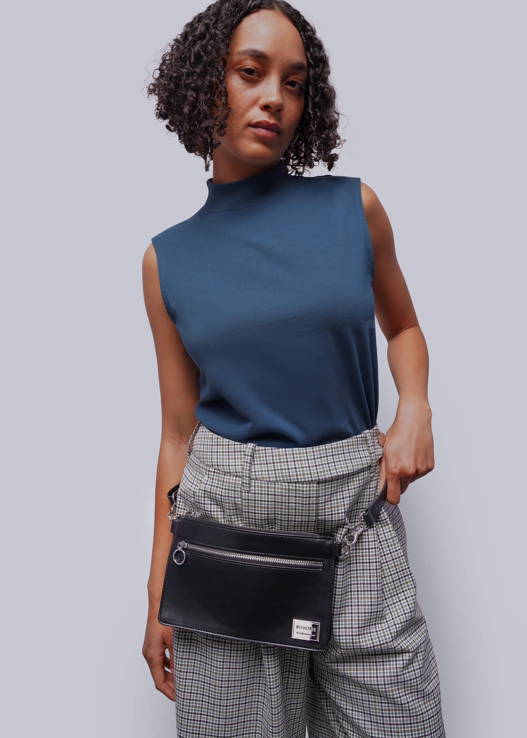 Woman wearing a blue sleeveless top and plaid pants with a black belt bag against a gray background