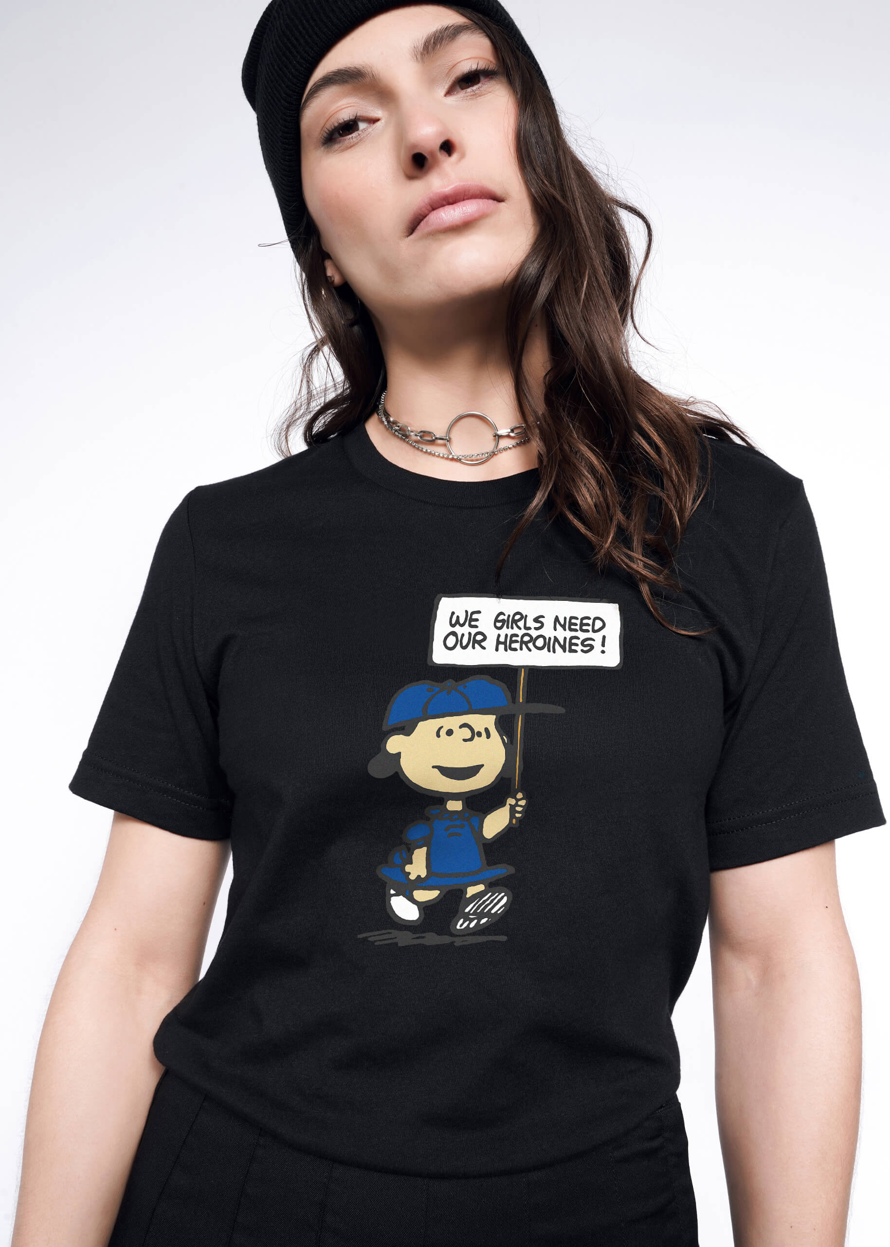 Person with long brown hair wears black beanie and The Peanuts x Wildfang Heroines Graphic Tee, which is a black tee that features a graphic of Lucy with a sign that says 
