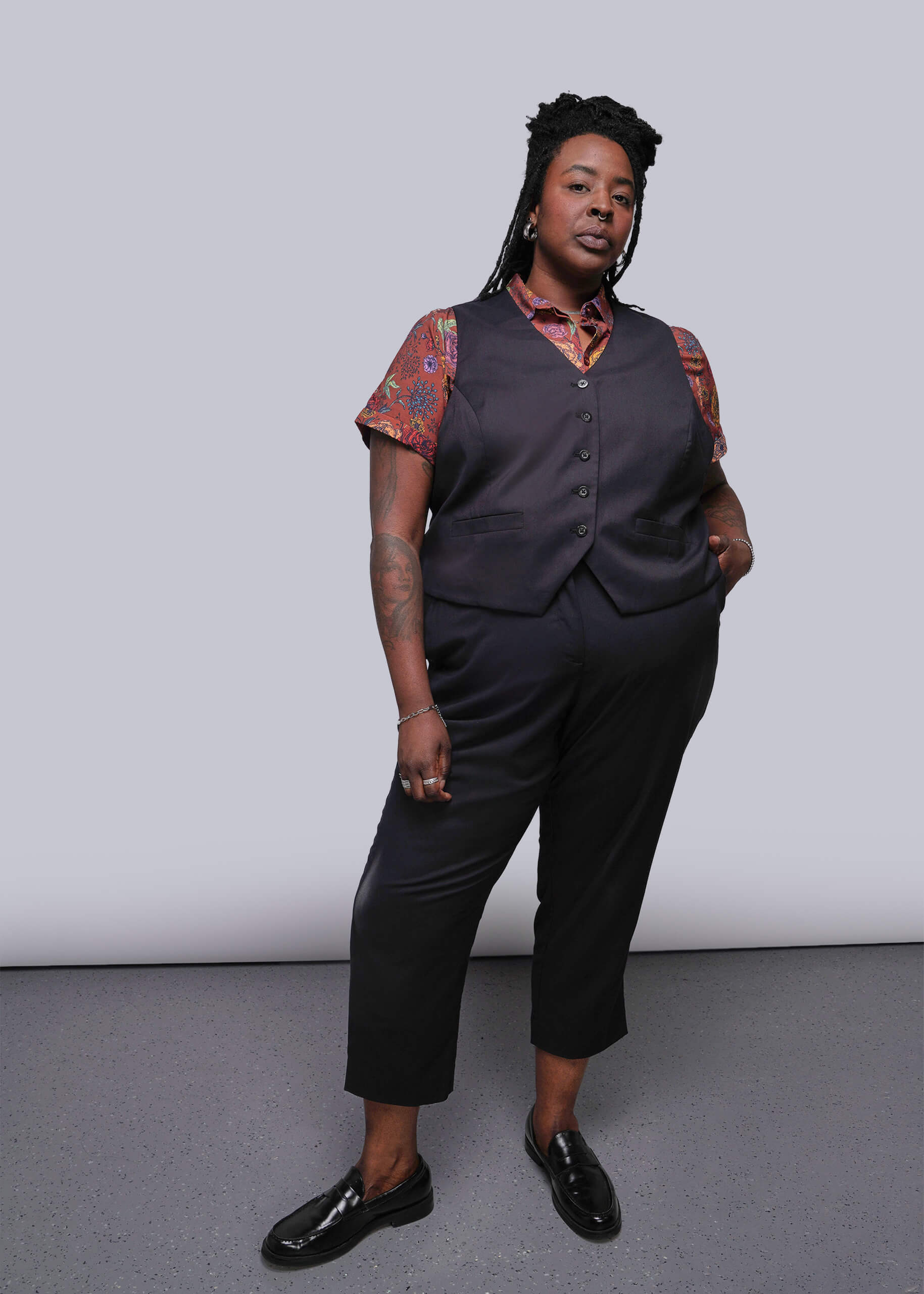 1X=Person wearing a dark vest over a patterned shirt with black pants against a plain background