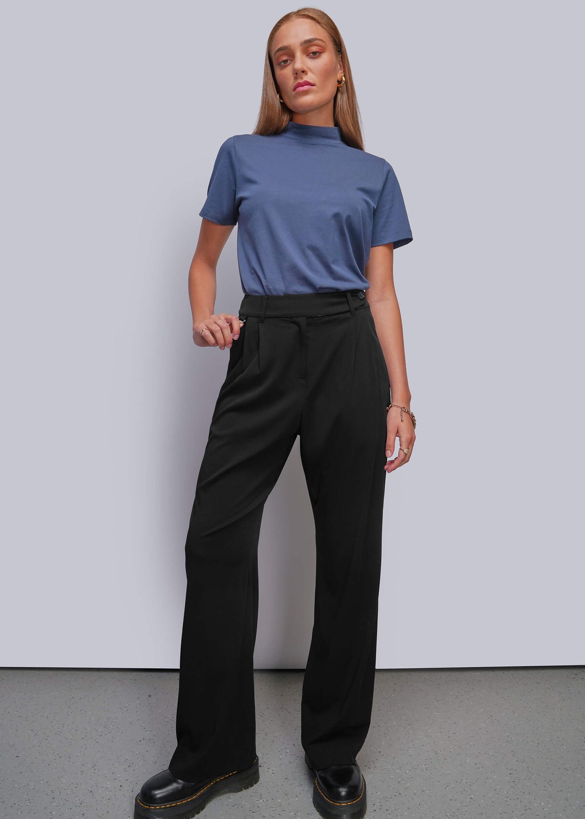 S=A woman modeling a blue short-sleeve mock neck top and black wide-leg trousers with black platform shoes, standing against a light gray studio wall.
