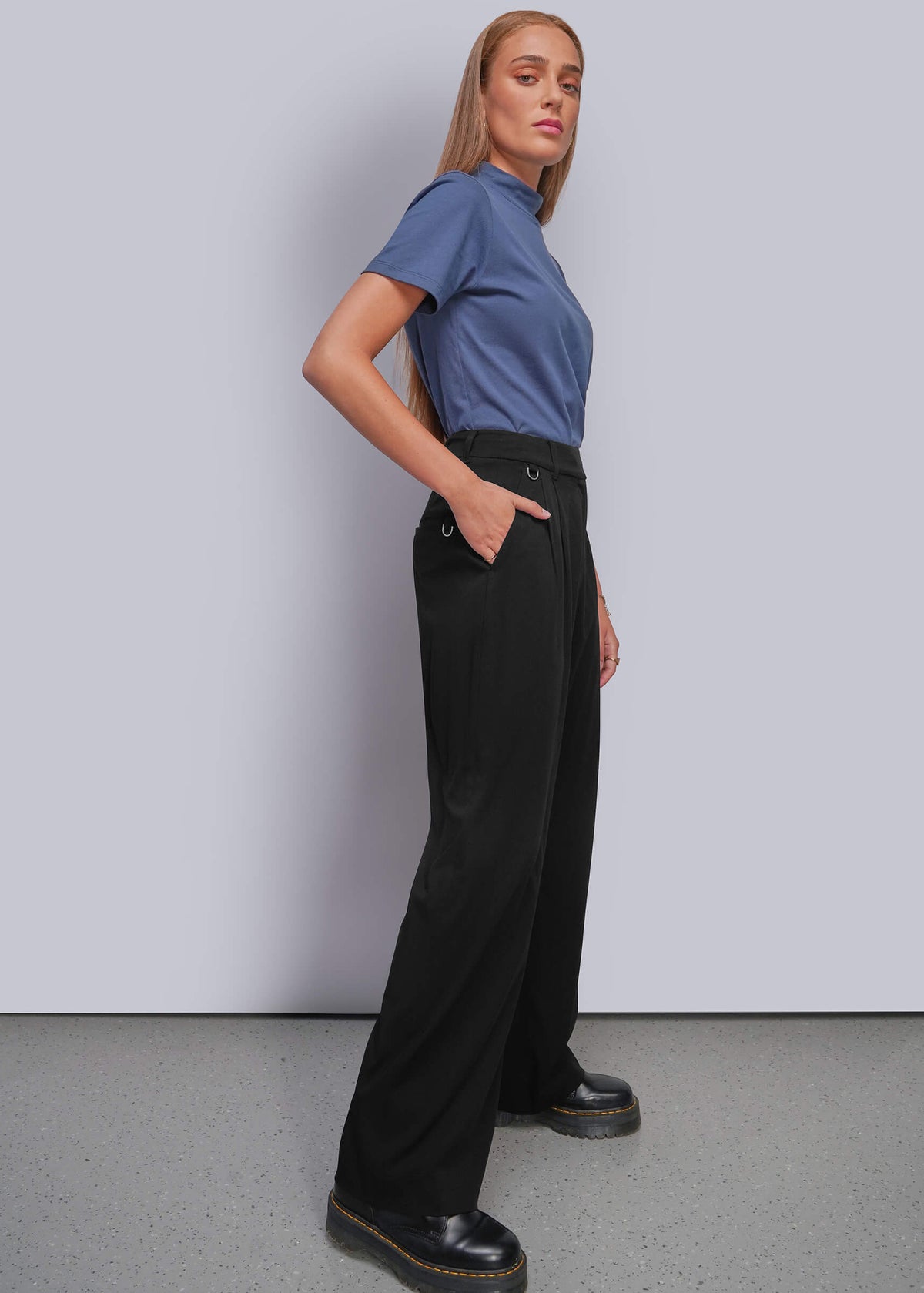 S=A woman modeling a blue short-sleeve t-shirt and black wide-leg trousers with black platform shoes, standing against a light gray studio wall.