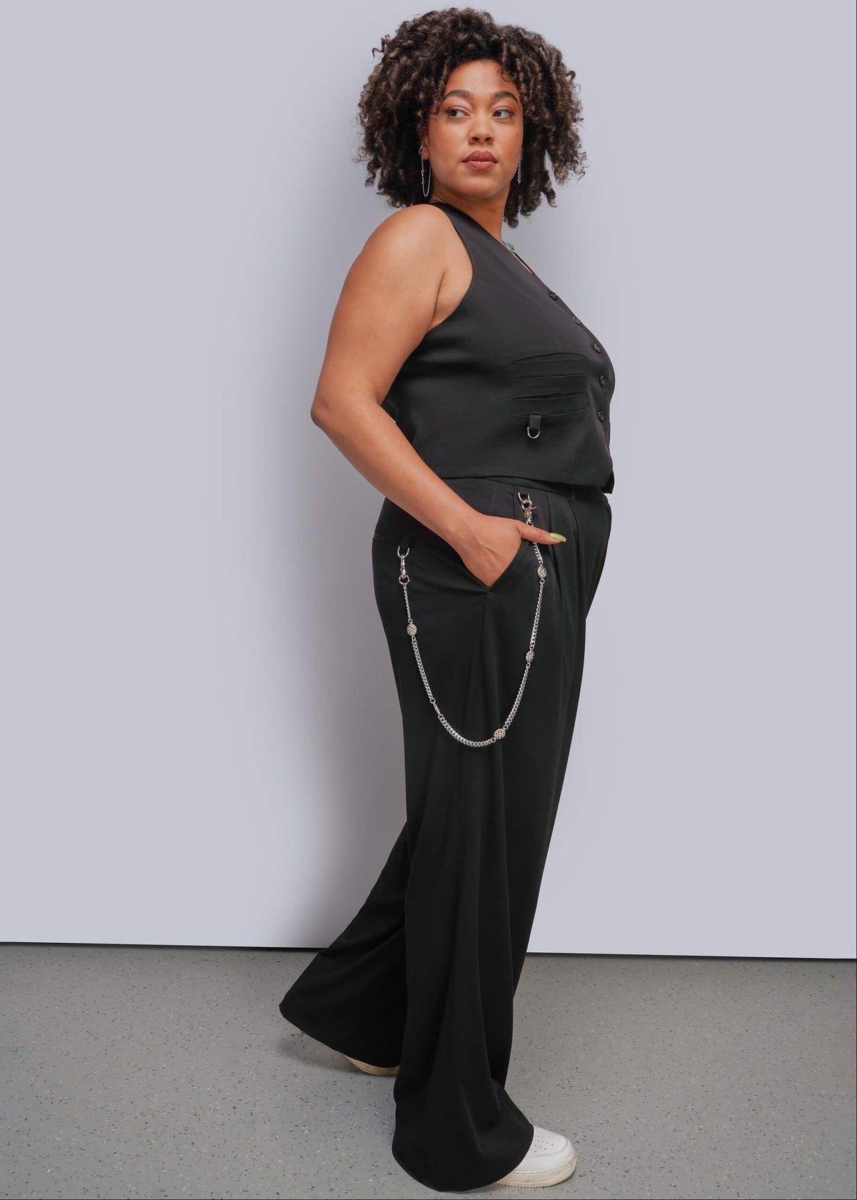 1X=A woman modeling a black sleeveless vest and matching wide-leg pants with a silver chain detail, standing against a light gray studio wall.