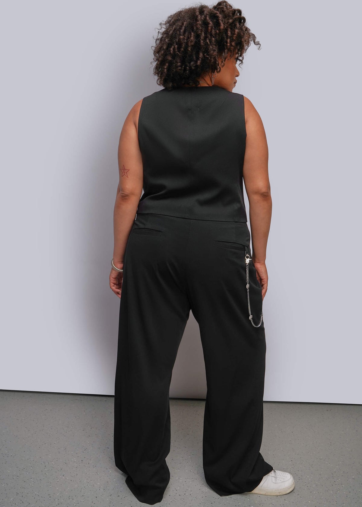 1X=A person shown from the back wearing a black sleeveless top and matching black wide-leg pants, standing against a plain light gray wall and gray floor.