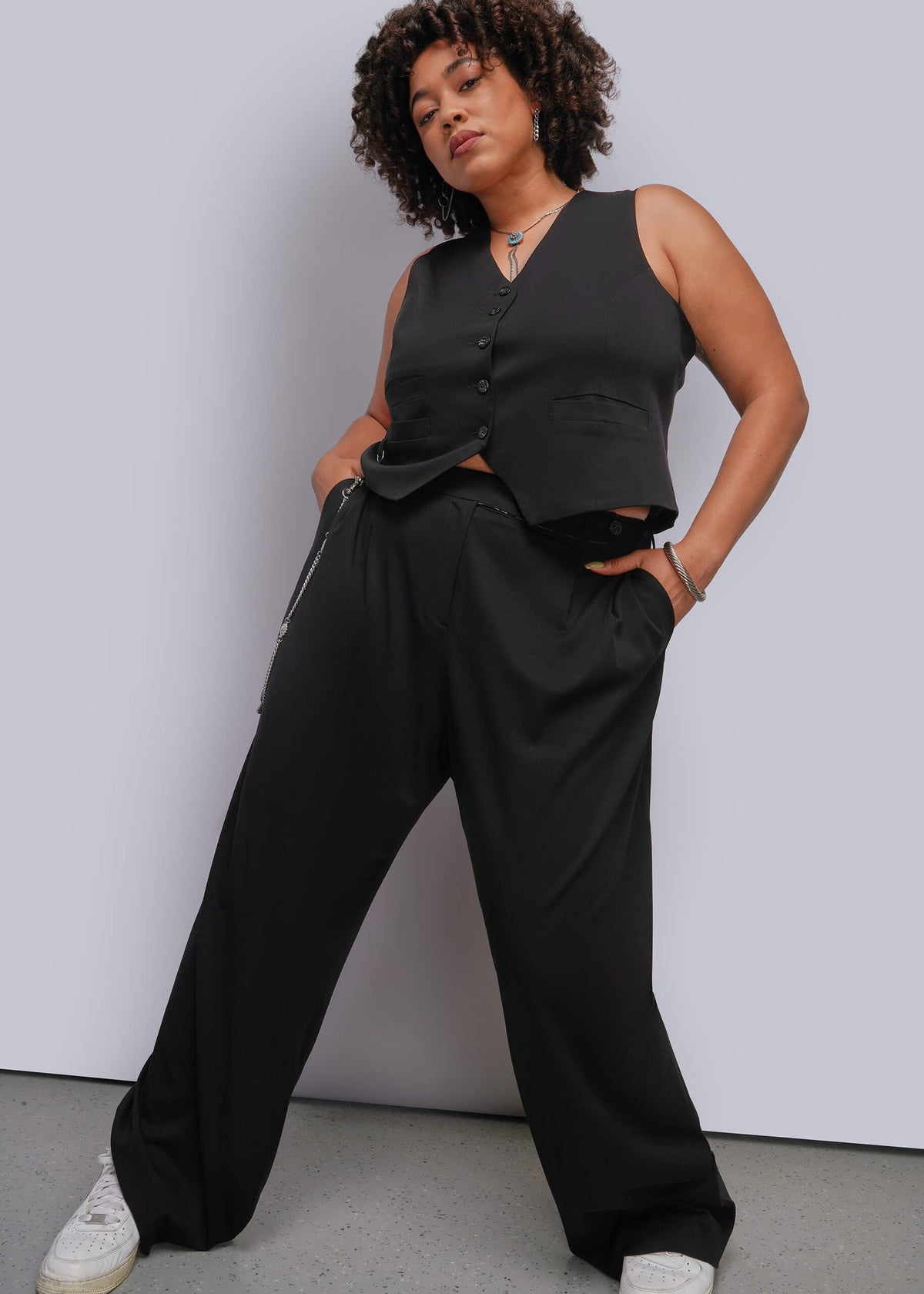 1X=A woman modeling a black sleeveless button-front vest and matching black wide-leg pants, paired with white sneakers, against a light gray studio wall.