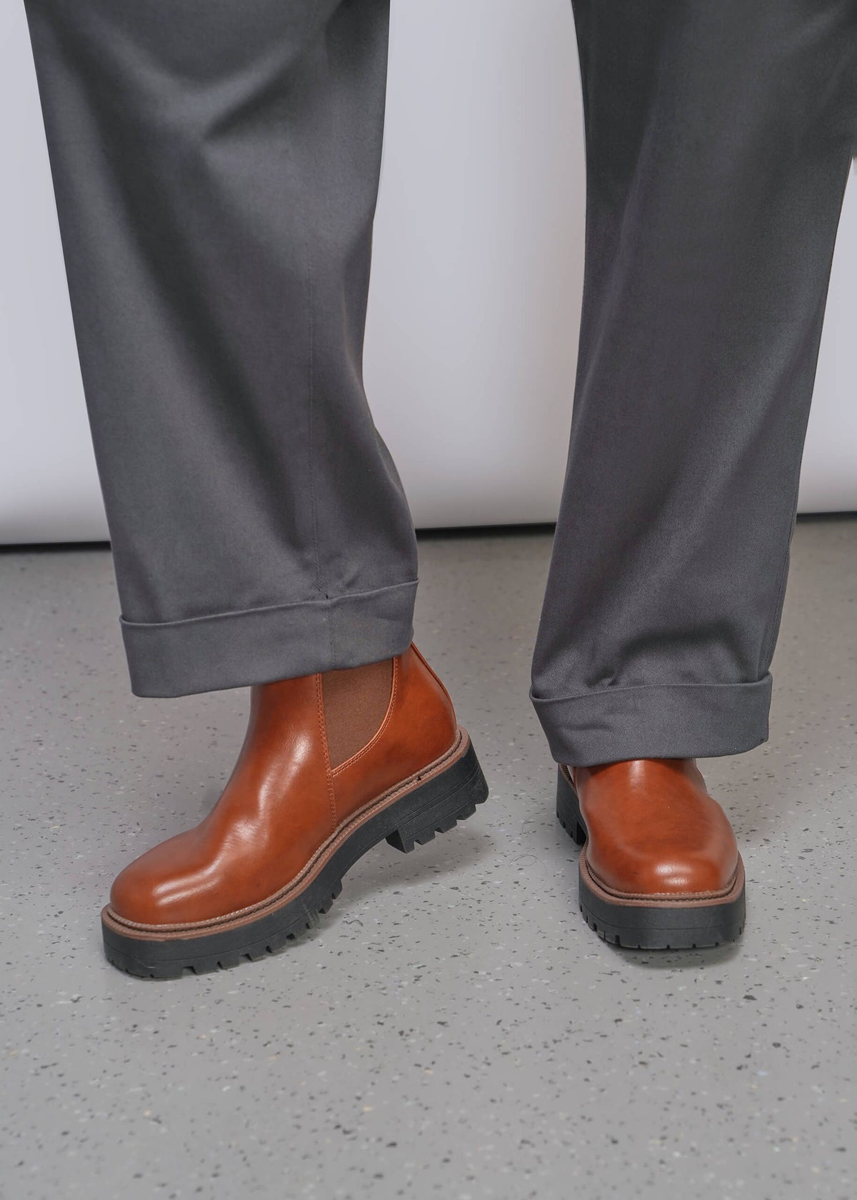 1X=Close-up of the pants part of a jumpsuit where the model is wearing brown leather Chelsea-style boots, standing on a speckled gray floor against a light gray wall.