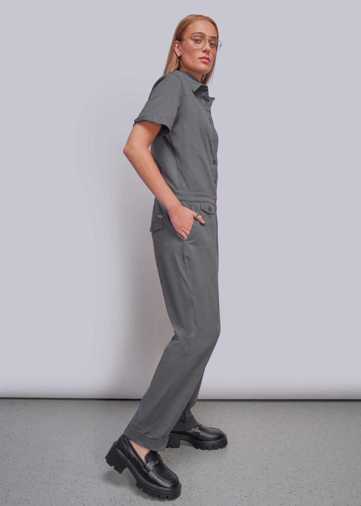 S=A woman modeling a gray short-sleeve collared jumpsuit with side pockets, paired with black loafers, standing in profile against a light gray studio background.