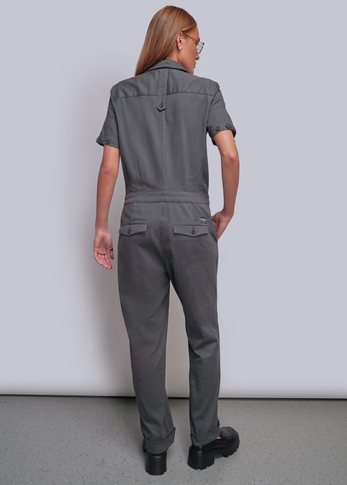 S=A woman viewed from the back wearing a dark gray short-sleeve jumpsuit with a collar and back pockets, standing against a plain light gray studio wall.