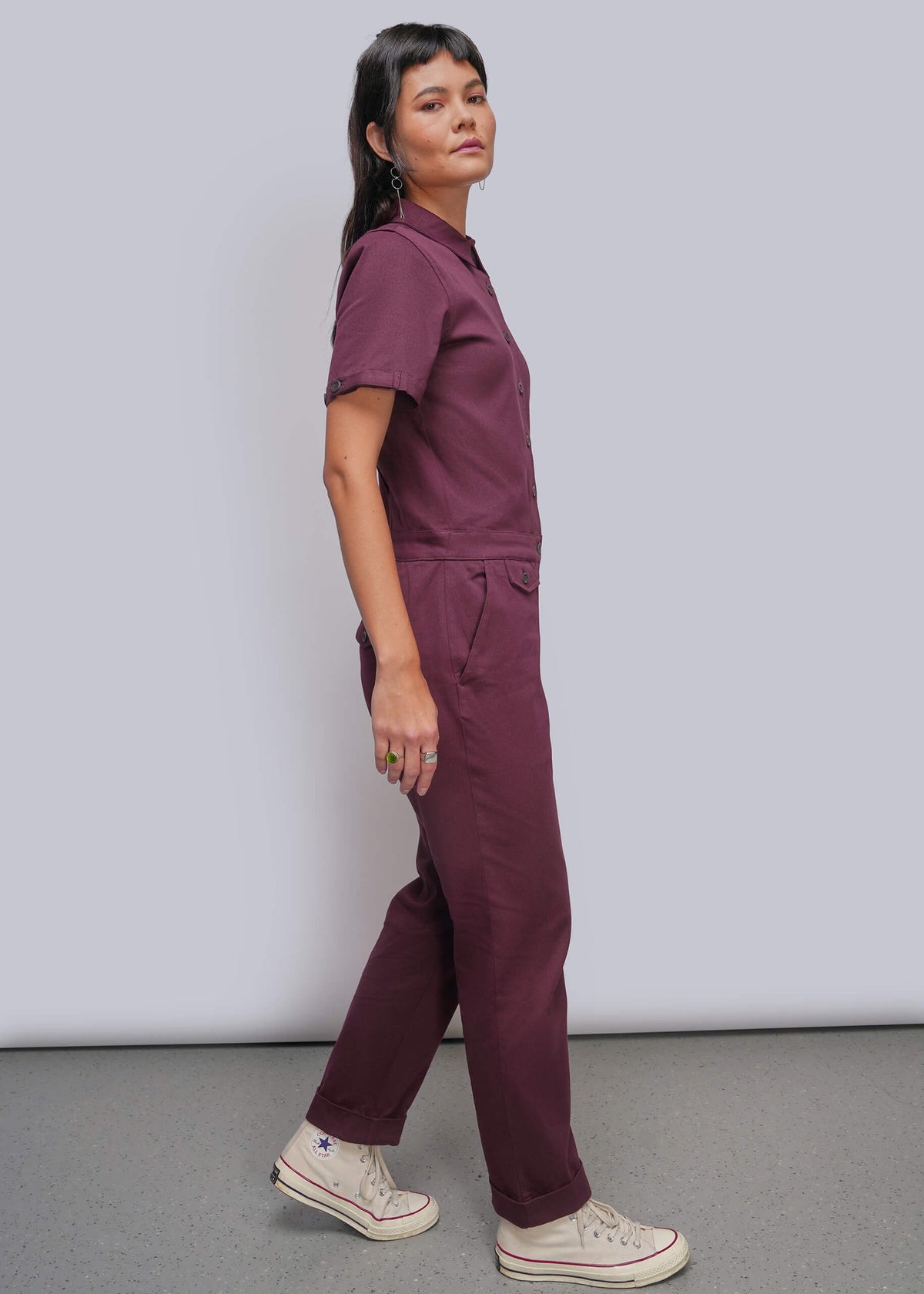 S=A person modeling a matching short-sleeve button-up jumpsuit in a solid burgundy shade, paired with off-white canvas sneakers, against a light gray studio background.