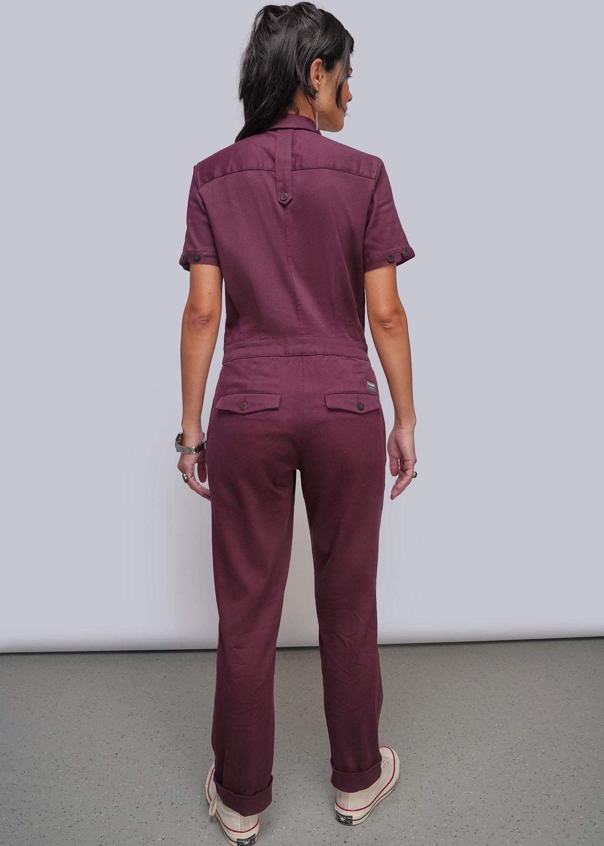 S=A woman viewed from the back wearing a short sleeved jumpsuit in a deep burgundy or plum shade, standing against a plain light gray studio wall.