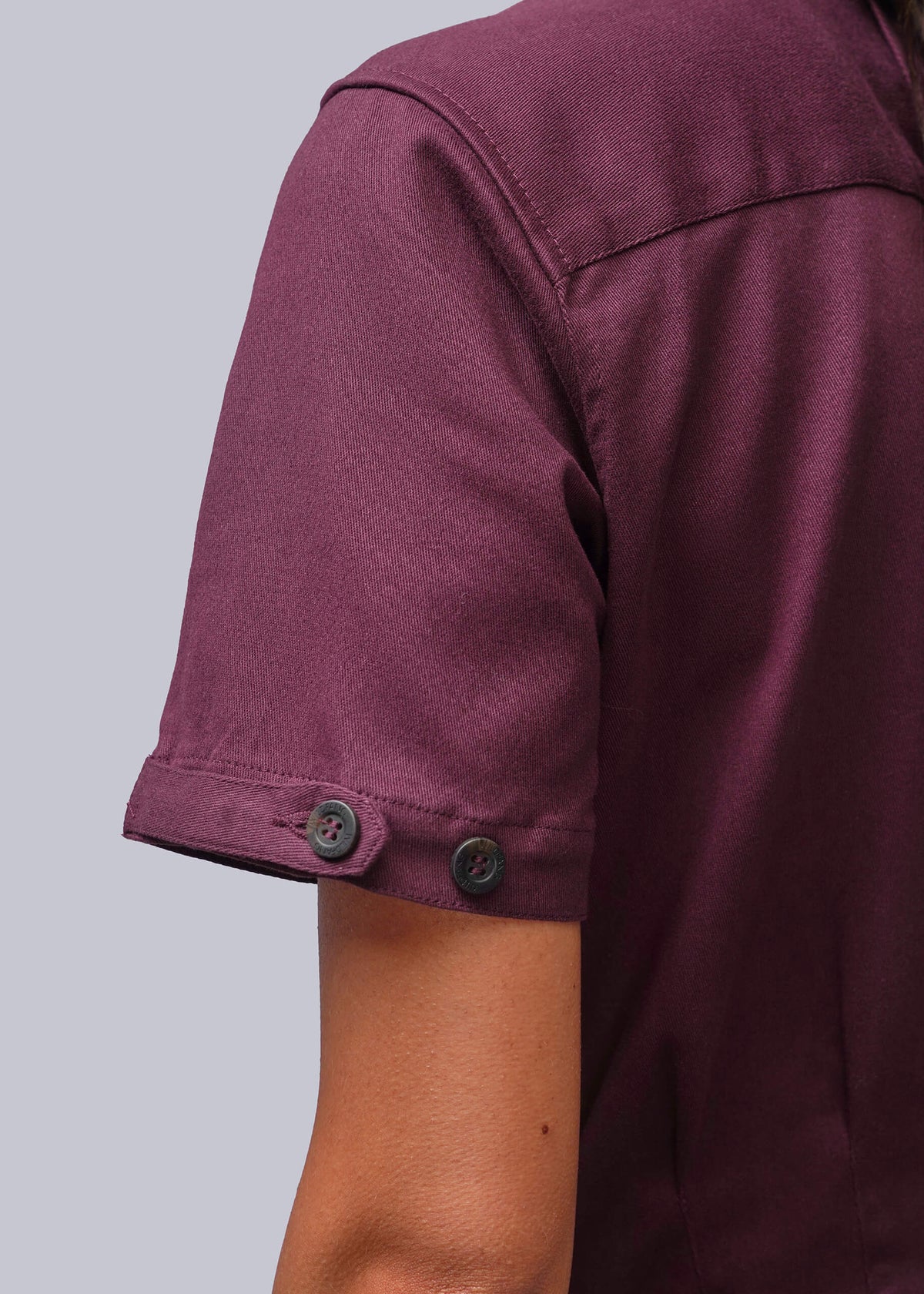 S=Close-up of a person wearing a short-sleeve jumpsuit in a deep plum or burgundy shade, showing the sleeve with two dark buttons and part of the arm against a light gray background.