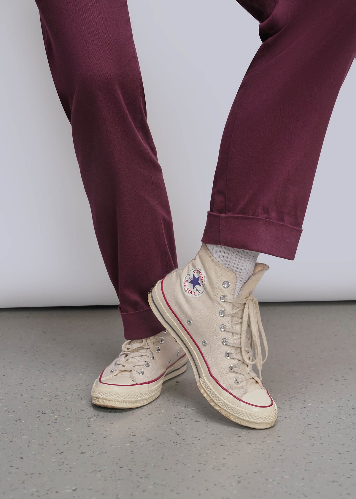 S=Close up of a person's burgundy jumpsuit pant cuffs and cream high-top canvas sneakers with red piping and a Converse logo, standing against a light gray wall and concrete floor.